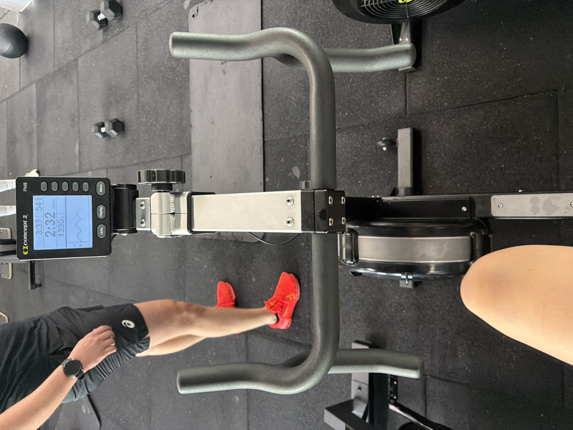A person is seated at a Concept2 rowing machine on a black rubber floor. The person is wearing black shorts, a black watch, and bright red running shoes, and the machine monitor shows a 500m split time of 2:32. The surroundings include a medicine ball and dumbbells, indicating a gym environment. The image does not contain any information to ascertain whether the location is in Sofia, Bulgaria, nor any contextual details to substantiate such a claim.