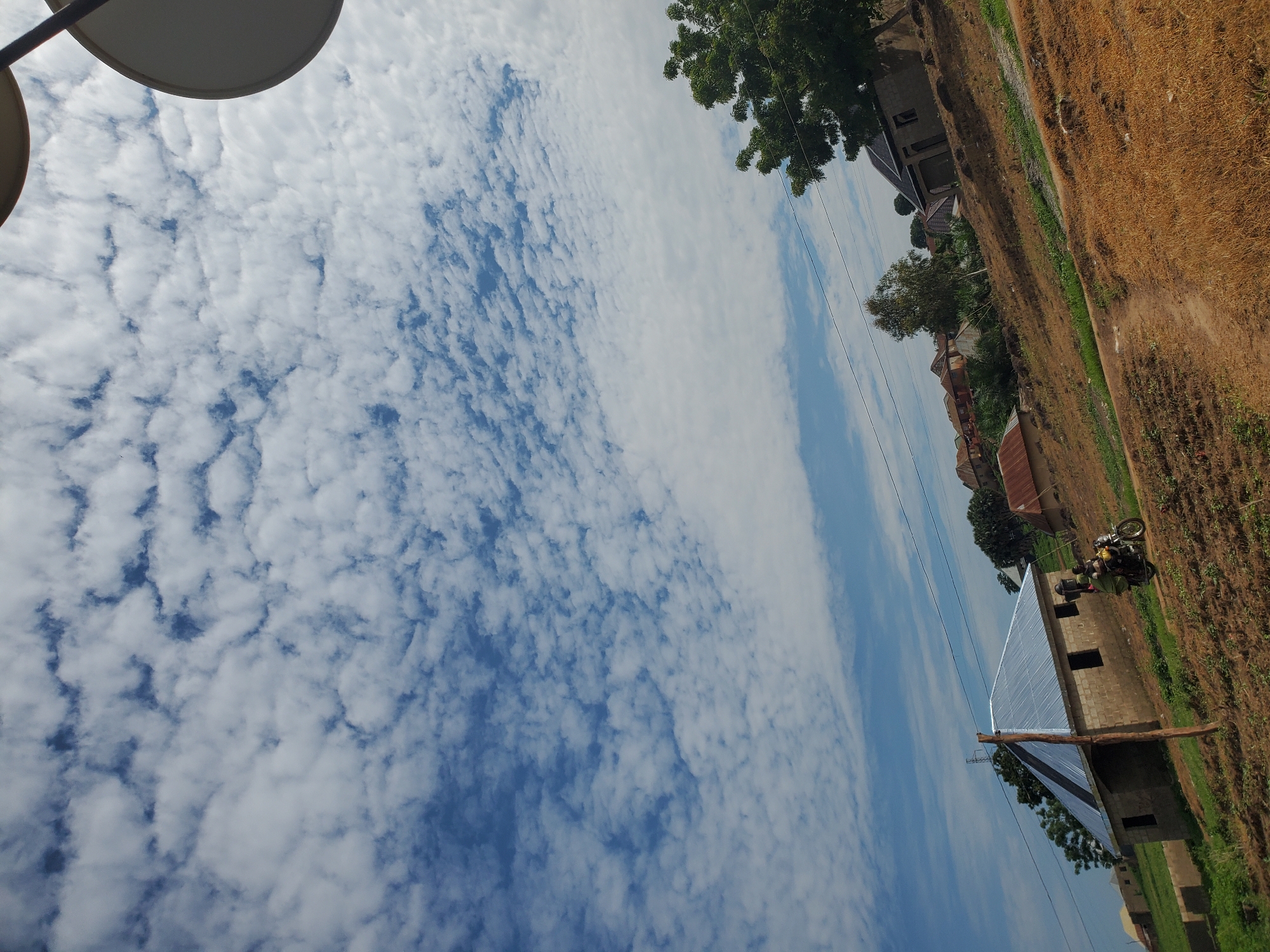 This image depicts a scene in the city of Winters, United States of America. The upper two-thirds of the vertical composition showcase a bright blue sky filled with numerous scattered, small white cumulus clouds, some forming distinct rows and patterns, suggesting an altocumulus or cirrocumulus formation. The clouds cover a significant portion of the visible sky, with patches of clear blue visible between them.
The lower third of the image presents a semi-rural landscape with a gently sloping, unpaved dirt path or road traversing from the foreground towards the midground. The ground cover consists primarily of dry, reddish-brown earth and sparse, dry grass, interspersed with patches of greener vegetation, particularly around the structures.
On the left side of the midground, a rectangular, partially constructed building with unfinished concrete block walls and a new, shiny, corrugated silver-colored metal roof is prominent. It features multiple rectangular openings serving as windows and possibly a door, lacking frames or glass. A raw wooden utility pole stands near this building. Further into the midground, additional residential structures with varying roof materials, some appearing to be corrugated metal or possibly thatched, are visible on higher ground.
In front of the partially constructed building, near the center of the image, a person dressed in dark clothing is riding a dark-colored motorcycle along the dirt path. A second person or a substantial item of cargo appears to be positioned on the rear of the motorcycle.
Several trees are scattered across the landscape. A prominent tree with dense green foliage occupies the right midground, partially obscuring the view of the sky and another building with a darker roof. Overhead utility lines are visible stretching across the midground, connected to poles. In the upper right corner, two circular, light-colored objects, consistent with satellite dishes, are partially cropped, with their dark mounting poles also visible against the sky.