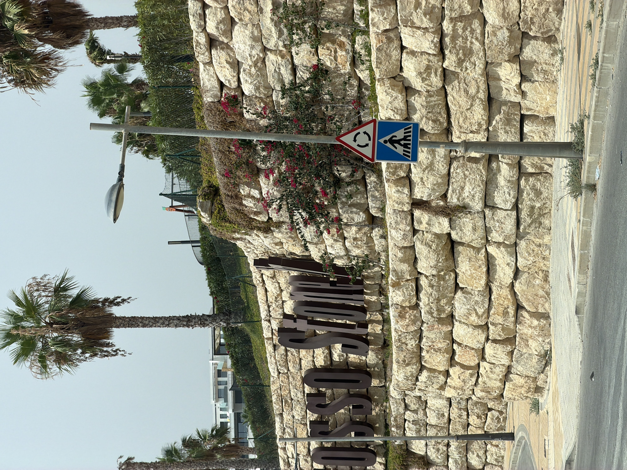 The image shows a large stone retaining wall in Finestrat, Spain, with the words "OSSO STONE" prominently displayed in large, dark letters on its surface. The wall is constructed from irregularly shaped, light-colored stones stacked to create a textured and rustic appearance.
At the top of the wall, several palm trees with their fronds are visible against a bright, overcast sky. The vegetation on and around the wall includes some flowering plants with small red blooms, as well as green vines and shrubs. In the background, modern white buildings with multiple stories can be seen, suggesting a residential or commercial development.
In the foreground, a lamppost stands beside the retaining wall, supporting a street lamp. Below the lamppost, two traffic signs are mounted: the upper sign is a triangular warning sign with a circular arrow indicating a roundabout, and the lower sign is a blue square sign with a white silhouette of a pedestrian crossing a crosswalk.
The area in front of the wall features a paved sidewalk and a road. The sidewalk has a curb separating it from the road, and sparse vegetation grows along the edges. The road surface is asphalt. The scene appears to be a public area, likely a street or entryway.