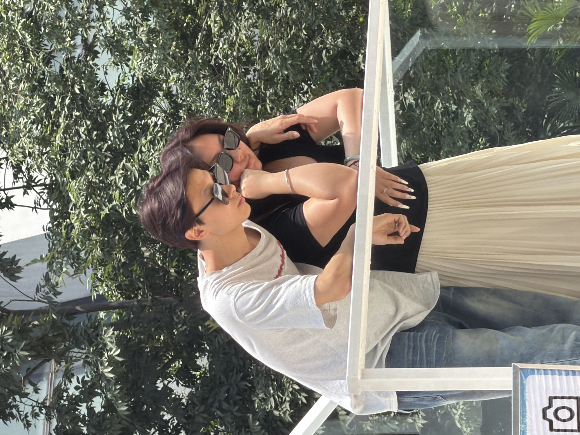 A young Asian couple is leaning on a white railing outdoors, with a lush green leafy tree canopy in the background. The male, on the left, wears a light gray t-shirt and blue jeans. The female, on the right, is wearing a black t-shirt and a long, beige pleated skirt. Both are wearing sunglasses. The male has his left arm around the female's shoulders. The female has her left arm around the male's waist, and her right hand is resting on her chin. A partial view of a blue and white sign with a camera icon is visible in the lower left corner. The location is Yuzhong District, China.