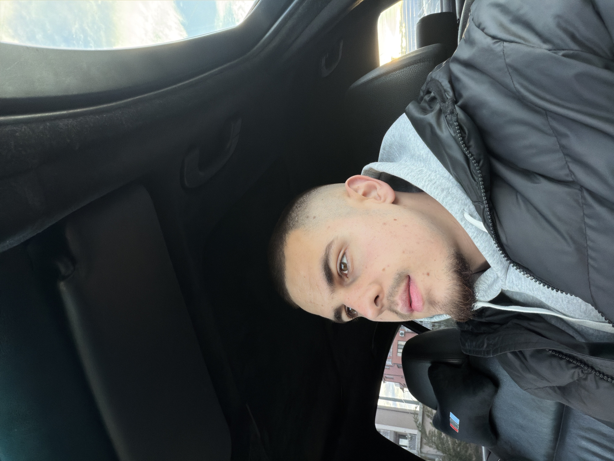 A young man sits inside a car, possibly in Plovdiv, Bulgaria given the general architecture and foliage outside the window. He's wearing a grey hoodie under a black puffy jacket. The interior of the car is primarily black. The window shows a glimpse of buildings and trees. The man has short hair and a short beard. The photo appears to have been taken from a low angle looking upwards.