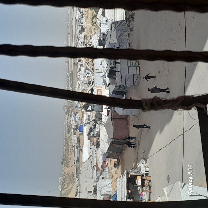 A large refugee camp is viewed from a window with a lattice. The camp consists of many small, makeshift tents and dwellings spread across a flat, arid landscape. Several individuals are walking along a dirt path that runs through the center of the camp. The scene is muted and dusty, and the horizon appears hazy.