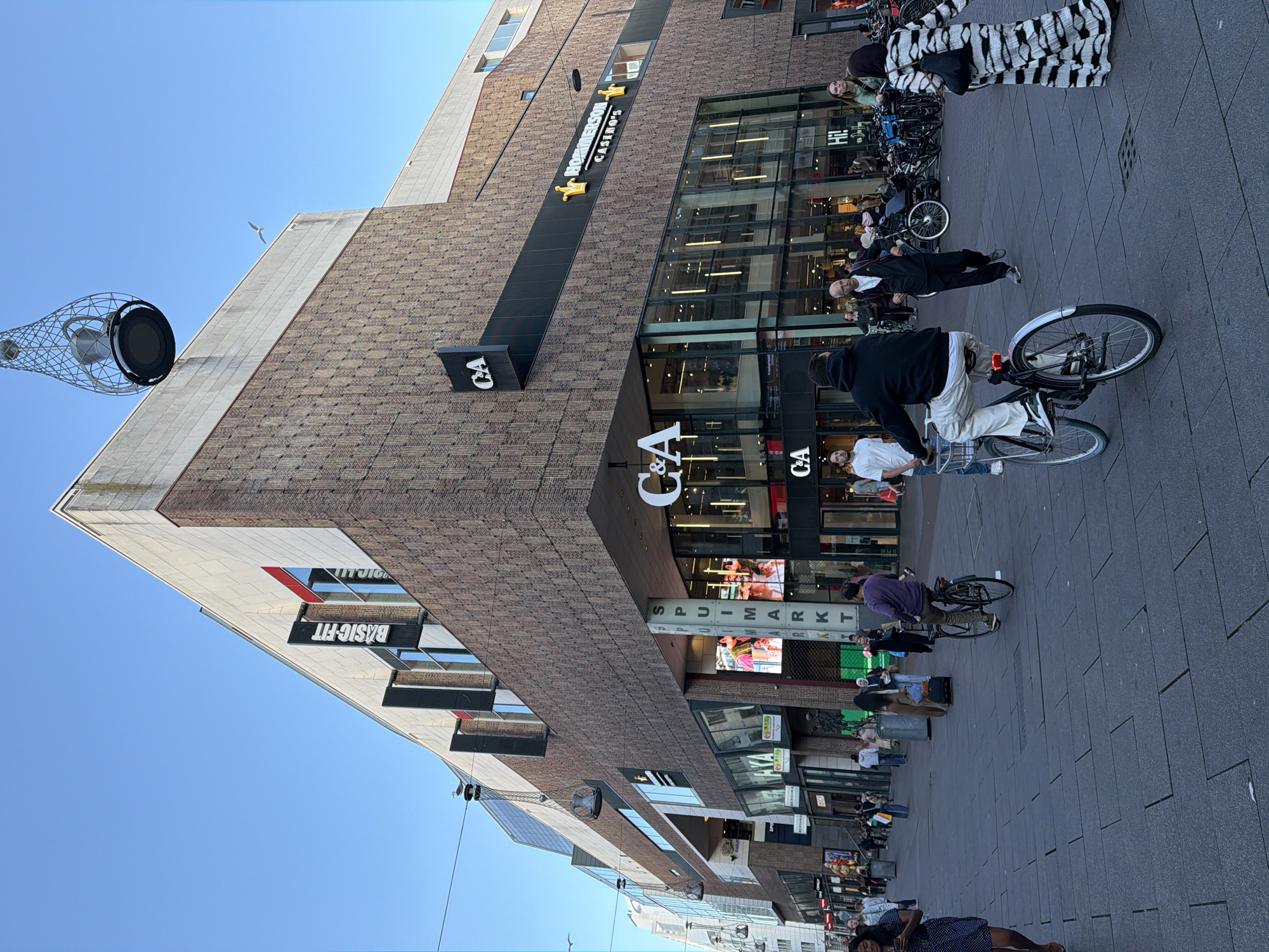 This is a street-level, medium-shot photograph taken in the city of The Hague, Netherlands, likely on a commercial street. The central focus is a large, modern building with a brick facade and a prominent C&A department store signage visible on its lower levels. The building also features "Basic-Fit" and "Hommerson Casino's" signs. The street is paved with dark gray square tiles and is occupied by pedestrians and cyclists.
In the foreground, a person rides a bicycle towards the viewer's right, wearing light-colored pants and a dark hoodie. To the right of the cyclist, another person is walking away from the viewer, wearing a distinctive black and white striped dress. Several other individuals are visible in the background, walking, cycling, or standing, with some bicycles parked to the right. The environment suggests a bustling urban shopping area with shops lining the street. The sky is clear and blue, indicating daylight. Above the building, an unusual, cage-like light fixture is suspended.