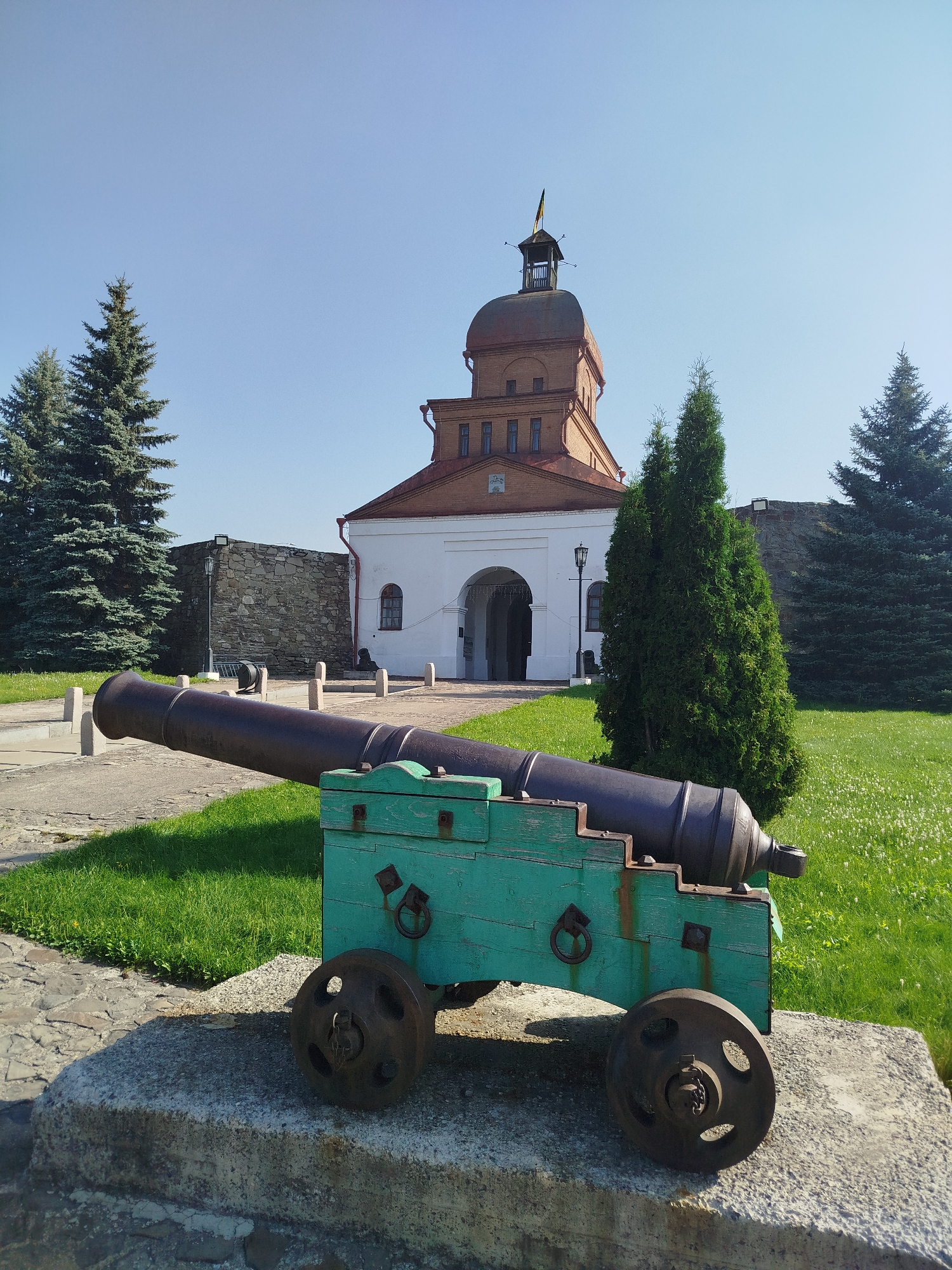 The image depicts a historical scene with a vintage cannon prominently displayed in the foreground. The cannon, mounted on a green wooden carriage with weathered metal wheels, rests on a stone foundation. Behind the cannon, a paved pathway leads to a large, white arched entryway of a brick building with a domed roof and a small tower topped with a flag. The building appears to be a historical landmark, possibly a fortress or a museum. The surroundings include lush green grass, scattered white flowers, and several tall evergreen trees. A stone wall extends from the building, suggesting it is part of a larger fortification. The sky is clear and blue, indicating a bright, sunny day. The scene is serene and appears to be a public space, possibly a park or a historical site within the city of Novokuznetsk, Russia, given its architectural style. No people are visible in the image.