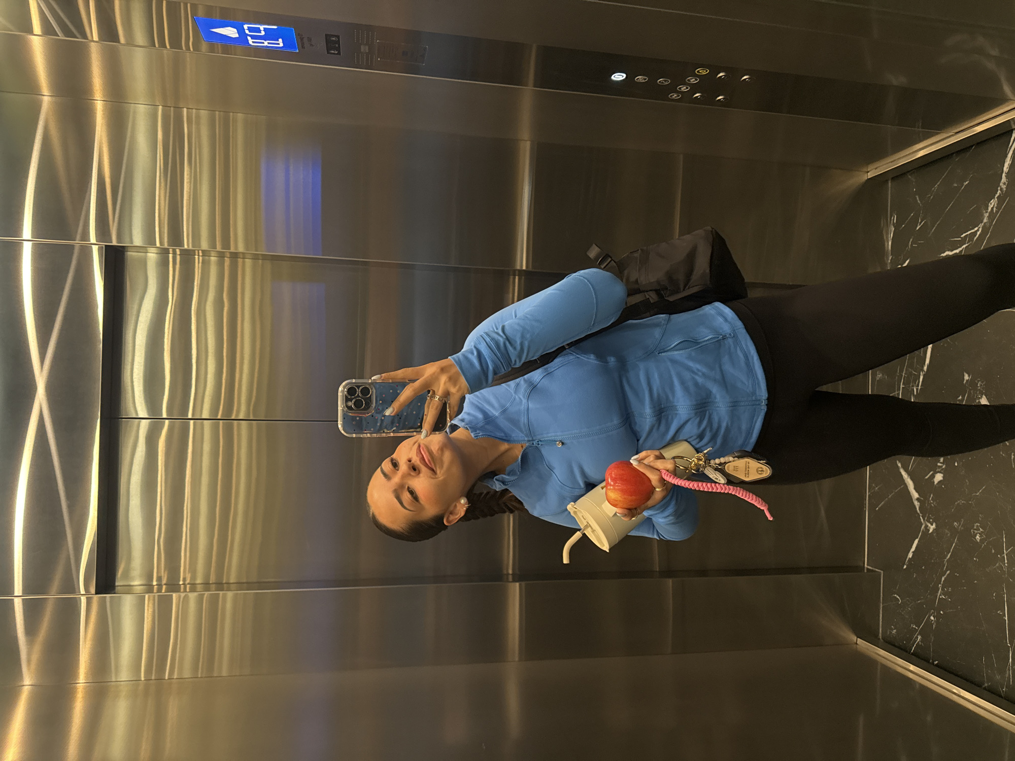 A woman stands in an elevator with stainless steel walls, holding a smartphone in her left hand to take a selfie. In her right hand, she holds a red apple, a white reusable coffee cup, and a keychain with keys attached to a pink woven strap. She is wearing a blue athletic jacket, black leggings, and has a black backpack slung over her shoulder. The elevator has a control panel with illuminated floor numbers and call buttons. The floor of the elevator is dark marble with white veining. The location is not indicated by visual cues in the image.