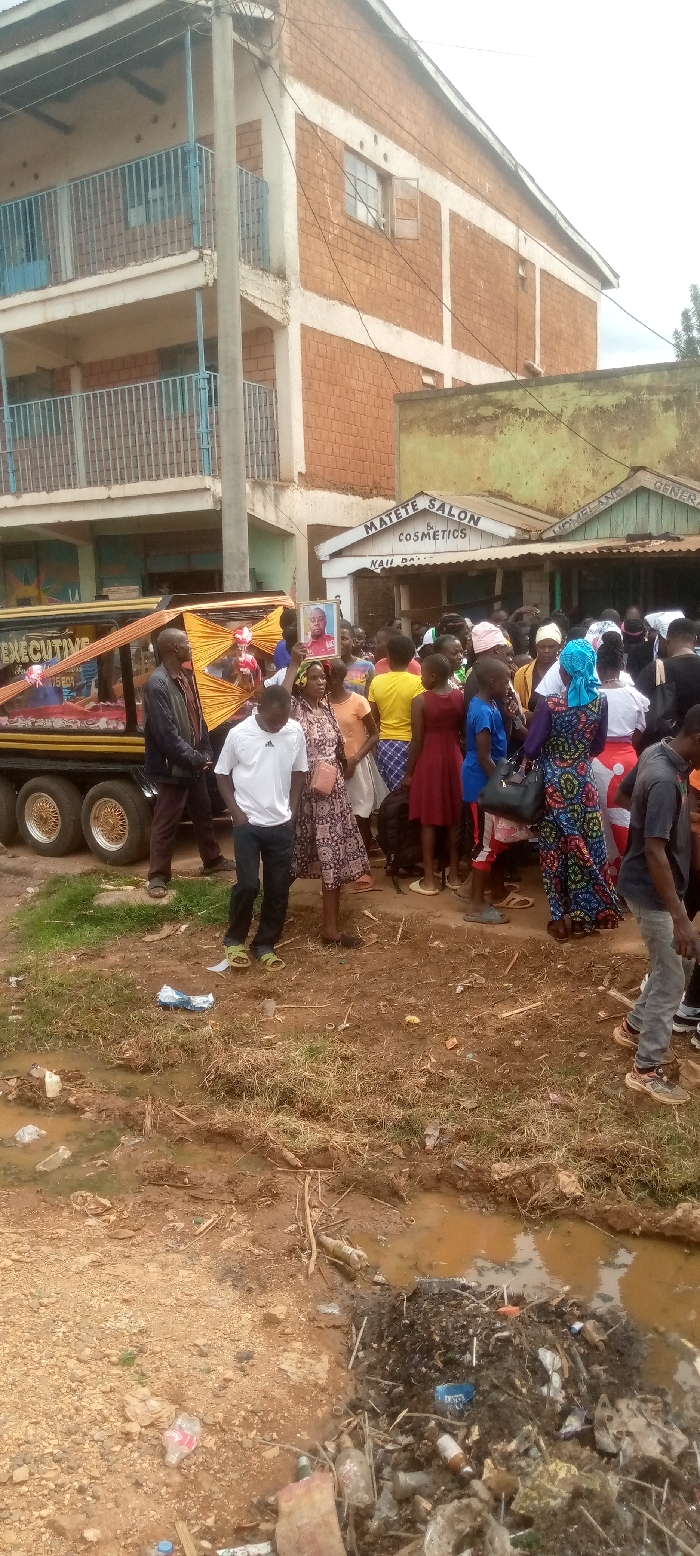 The scene takes place in a small town in Kivaywa, Kenya. A group of approximately 30 people are gathered around a hearse. The hearse is decorated with yellow and gold fabric. It is parked in front of a brick building that has a sign for "MATETE SALON & COSMETICS" above the entrance. The people are dressed in a variety of casual clothing and are talking to each other. Some of them are holding flowers and are walking behind the hearse. The hearse is facing a dirt road. The road is filled with garbage and is lined by a small, shallow ditch filled with murky water. The ground is dirt and grass. The sky is partly cloudy.