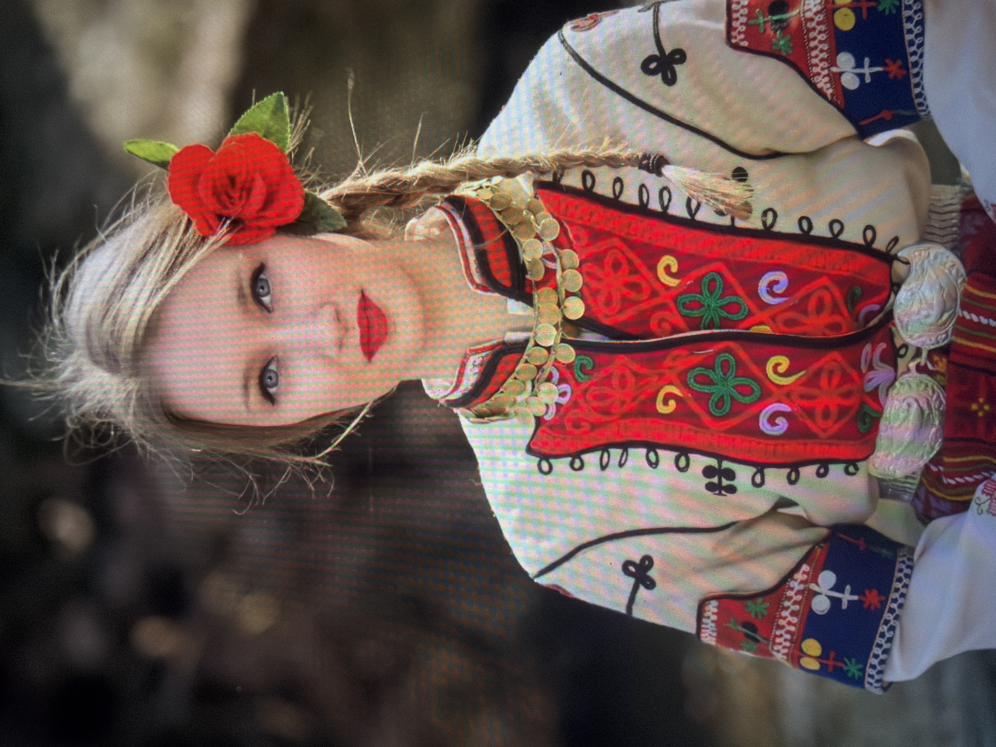 A young woman with fair skin, light blonde hair, and blue eyes is depicted in a medium shot, looking directly forward. She wears red lipstick and has a bright red artificial rose with two green leaves pinned to the right side of her head. Her hair is primarily styled in a long braid draped over her right shoulder, with several loose strands framing her face.
She is dressed in a traditional Bulgarian folk costume. This includes a white long-sleeved blouse, over which she wears a vibrant red sleeveless vest or bodice. The vest is richly embroidered with multi-colored patterns, primarily in green, yellow, and light blue floral or abstract motifs, and features black looped trim along its edges and down the front opening. Around her neck, she wears a multi-strand necklace composed of numerous small, gold-colored coins. The sleeves of her white blouse are adorned with distinct blue embroidered bands, which contain geometric and abstract symbolic patterns in white, yellow, and red. A patterned sash or belt, secured by an ornate, silver-toned buckle or clasp, is visible around her waist.
The background is dark, out of focus, and indistinct, suggesting an outdoor natural environment with diffused light sources creating blurred bokeh effects. The overall image exhibits visible pixelation, indicative of a photograph of a digital screen. The scene is located in Shumen, Bulgaria.