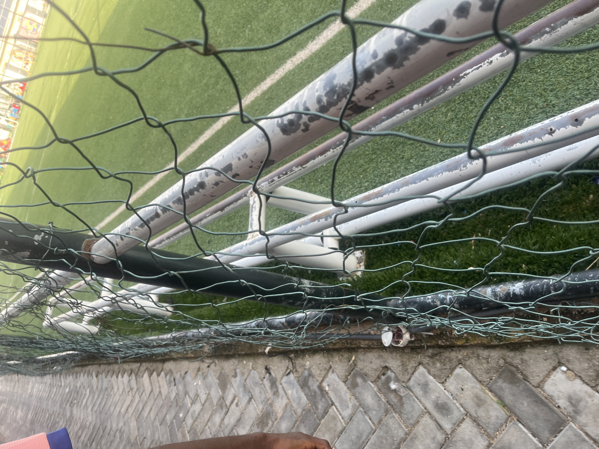 This is a close-up, low-angle shot of a chain-link fence in front of a soccer field. The fence is green and appears somewhat worn, with some rust visible on the metal poles it's attached to. Beyond the fence, the green artificial turf of a soccer field is visible, marked with white lines. The sun is shining, casting a bright light on the field, suggesting it is daytime. In the foreground, a person's hand is visible reaching towards the fence. The ground in the immediate foreground consists of patterned grey paving stones. The overall scene suggests an outdoor sports facility in Jalingo, Nigeria, likely a soccer pitch. There is no discernible text in the image. The presence of the fence and the stadium lighting (partially visible in the top left) indicates a public sports venue. The image is oriented vertically, giving a sense of looking up at the fence and the field.