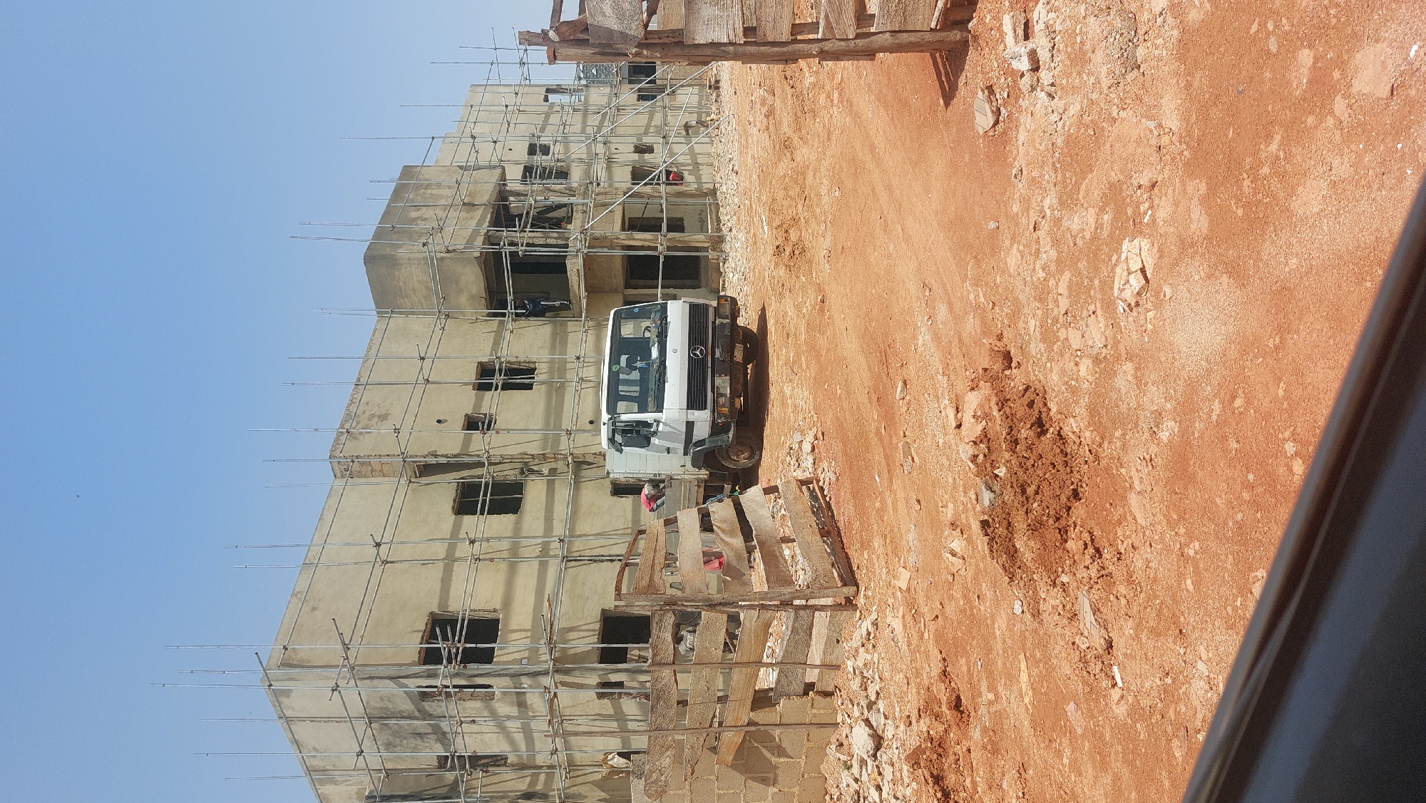 This image captures a construction site in Gura Top, Nigeria, under a clear, bright sky, suggesting a sunny day. The primary subject is a multi-story building under construction, encased in scaffolding. The building appears to be in an unfinished state, with visible concrete walls and openings where windows and doors will be.
In the foreground, a white flatbed truck is parked on a dirt road. The truck has a Mercedes-Benz logo on its grille. Scattered around the truck and on the road are piles of reddish-brown earth and debris, indicative of ongoing construction work. Wooden structures, possibly temporary supports or scaffolding elements, are also visible in the foreground.
There are a few individuals present at the site. One person in a red shirt can be seen near the truck, possibly a construction worker. Another figure is partially visible on the scaffolding of the building, also likely engaged in construction activities. The scene conveys a sense of active labor and progress on the building project. The overall impression is a typical day at a construction site in a developing area, with a focus on infrastructure development. No visible text is present in the image.