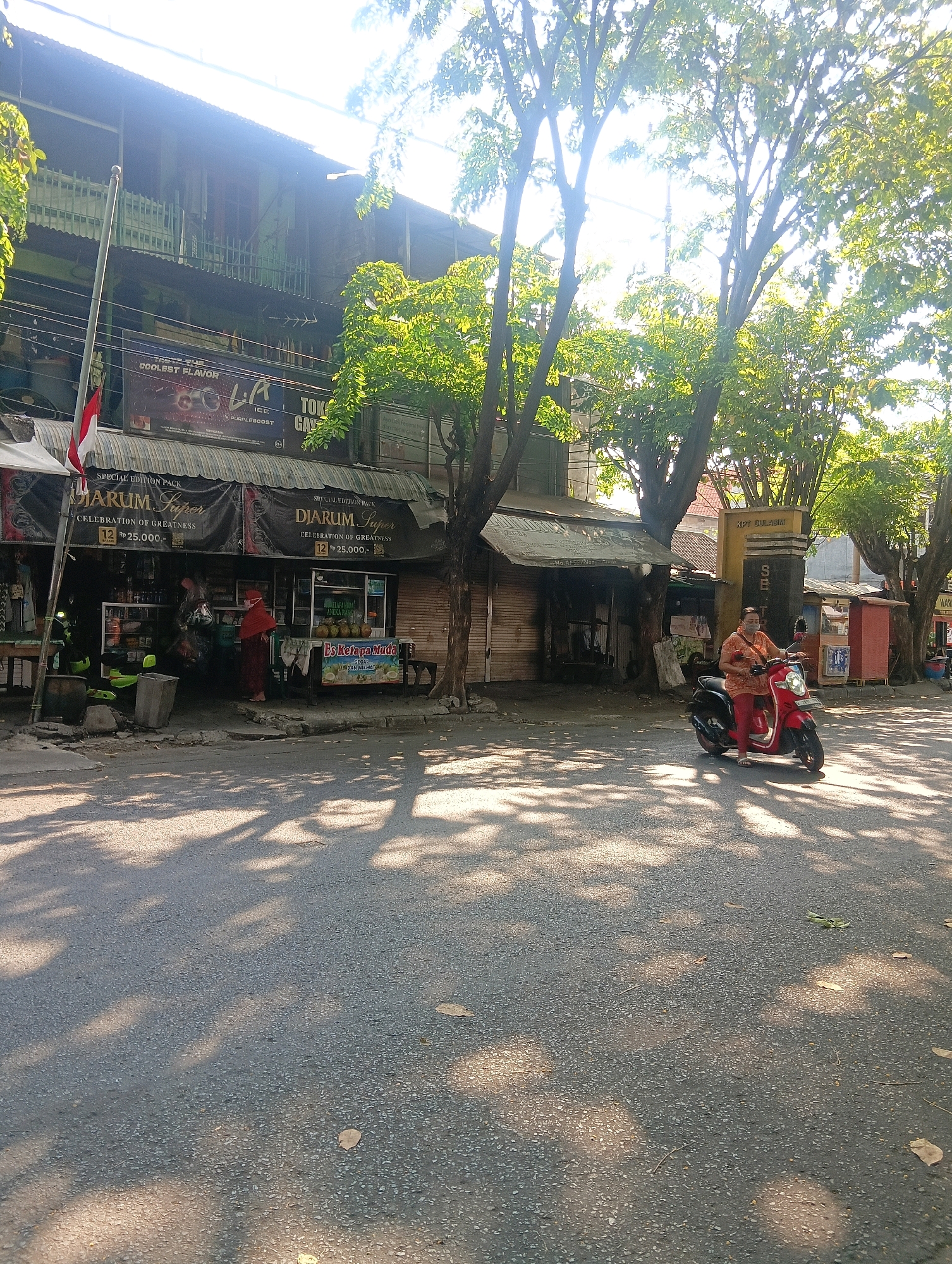 The image captures a street scene in Gapurosukolilo, Indonesia, on a bright, sunny day. The foreground is dominated by an asphalt road dappled with shadows cast by overhead trees. A few fallen leaves are scattered on the road surface. On the left side of the image, a building with multiple stories features balconies and a dark-colored awning above the ground floor. A flagpole with the Indonesian flag is positioned in front of this building. Signs indicate businesses, including one advertising "DIARUM Super" and "CELEBRATION OF GREATNESS" with prices in Indonesian Rupiah. A person in a red head covering and outfit stands near the entrance of a shop.
To the right, a woman wearing an orange top and red pants rides a red scooter. She is wearing a face mask. Behind her, a yellow pillar with dark lettering is visible, along with some smaller stalls or kiosks. The background shows more trees and buildings, suggesting a commercial or residential area. The overall impression is of a typical street in a town or city with active businesses and pedestrian and vehicular traffic.