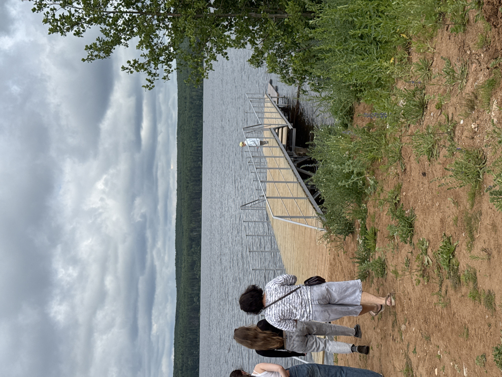The image depicts a natural landscape scene with a body of water, a wooden pier, and a group of people. The sky is overcast with a thick layer of clouds. On the left side of the image, three individuals are visible from behind, walking on a sandy path towards a wooden pier. One person is wearing a white top and blue jeans. Another person is wearing a black top and light-colored jeans. The third person is wearing a white and black striped shirt and light-colored shorts. On the pier, a fourth person is standing, facing away from the camera, wearing a light-colored top and shorts, and a yellow hat. The pier extends into a wide, calm body of water, likely a lake or reservoir. The opposite shore is lined with a dense forest of green trees. Lush green vegetation and some scattered wildflowers grow along the sandy embankment in the foreground. The scene appears to be a recreational area.