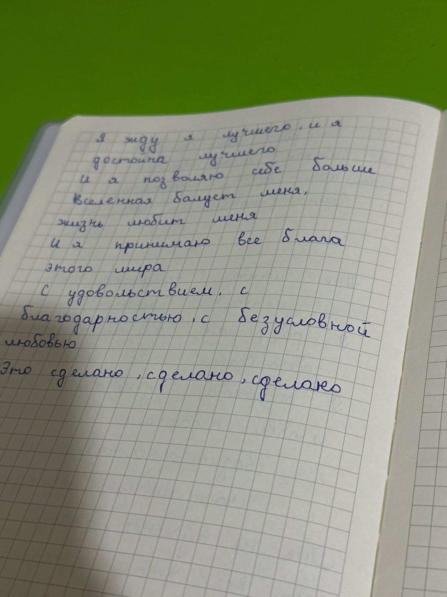 The image shows a handwritten poem on a page of a squared notebook. The poem is written in blue ink. The text of the poem reads: "Я жду я лучшего, и я достойна лучшего и я позволяю себе больше Вселенная балует меня, жизнь любит меня и я принимаю все блага этого мира С удовольствием, благодарностью, с безусловной любовью Это сделано, сделано, сделано". The notebook is open, with the right page visible and slightly angled. A portion of a green surface is visible in the upper left corner of the image, suggesting the notebook is placed on a green surface, possibly a table or desk. There are no people or other identifiable objects in the scene. The image does not contain any information about the location being Salar, Uzbekistan.