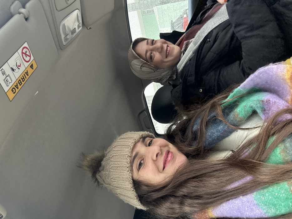 The image displays two adult females inside a vehicle, likely a car, captured in a selfie. The vehicle's interior features a grey headliner, a sun visor on the passenger side with a yellow "AIRBAG" warning label, and an overhead console light fixture. The individual on the left, positioned in the foreground, has long brown hair and wears a light beige ribbed knitted beanie with a brown faux fur pom-pom. Her lips are glossy pink, and she is smiling directly at the camera. She wears a multi-colored chunky knit scarf with prominent bands of purple, green, and blue, over a white garment. The individual on the right, positioned in the background, wears a light brown patterned headscarf (hijab), a black quilted jacket with a fur-lined collar, and a grey and reddish-patterned scarf. She is also smiling towards the camera. Through the vehicle's window, an overcast or rainy exterior is suggested by blurred reflections, with a red vehicle partially visible in the distance. While the image is associated with Meknes, Morocco, no specific landmarks are visible to pinpoint the exact location within the city.