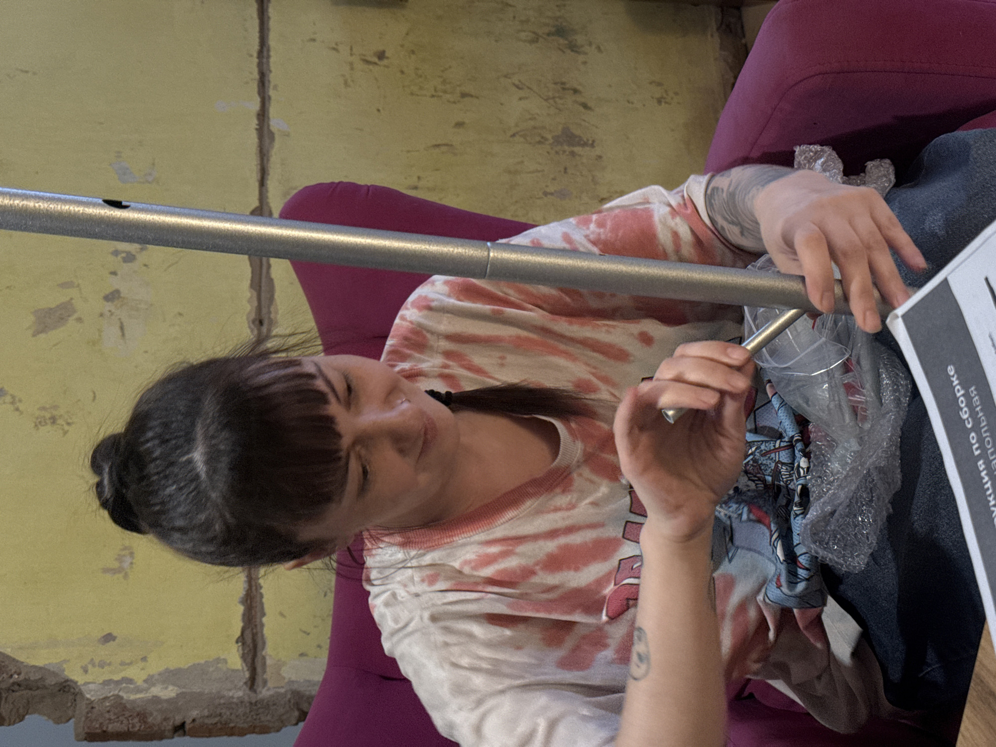 A woman is seated indoors, assembling a metal frame. She has dark hair pulled back in a bun, with bangs. She wears a pink and white tie-dye shirt. Her arms are tattooed. She sits on a pink chair. To her right is a set of instructions in Russian and packaged components. The room has yellow walls with visible aging and damage. This photo was taken in Votkinsk, Russia.