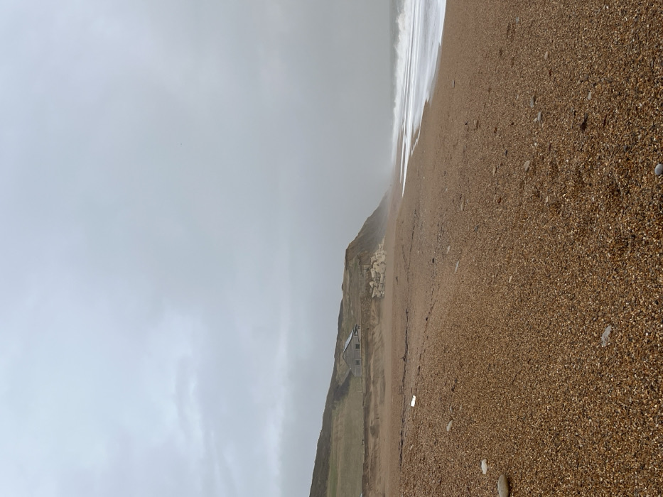 The image displays a coastal scene in Swyre, United Kingdom, under an overcast sky. The foreground is dominated by a broad pebble and shingle beach, exhibiting a reddish-brown hue with scattered lighter-colored stones and visible footprints. A single white piece of debris rests on the shingle in the mid-left. To the left, a gently sloping grassy hill transitions into an eroding cliff face of lighter brown earth. Perched on the lower slope of this hill, near the cliff edge, is a weathered, light grey or brownish-grey building with a gabled roof. A cluster of large, pale, angular rocks appears at the base of the cliff adjacent to the building. To the right, the sea features breaking waves with distinct white foam receding onto the shingle. The ocean surface appears grey under the uniformly light grey, cloud-covered sky, which extends across the entire upper portion of the frame. No individuals are present in the scene.