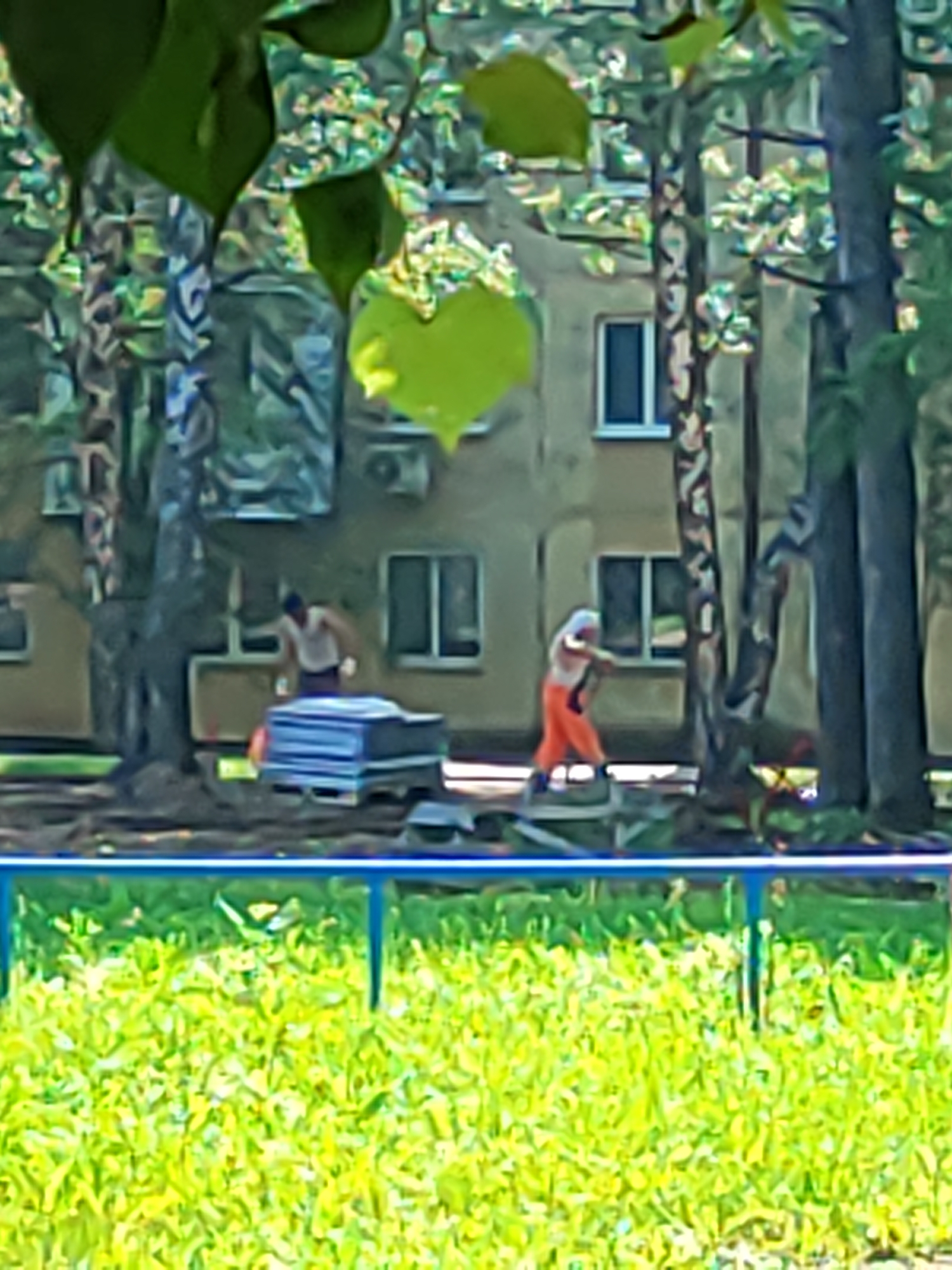 The image shows an outdoor scene with a building in the background and a grassy area in the foreground. Two individuals are visible in the mid-ground. The person on the left, wearing a white tank top and dark shorts, appears to be leaning over a stack of blue and white materials. The person on the right is wearing bright orange pants and a light-colored top and is actively engaged in some activity, possibly moving materials. They are positioned near the building, which has several windows. Trees with foliage are present between the building and the people, and more trees are visible to the right. The foreground features a bright green grassy area bordered by a blue railing. The overall impression is that of a construction or renovation site. Without additional information, the specific location within Ufa, Russia, cannot be determined.
