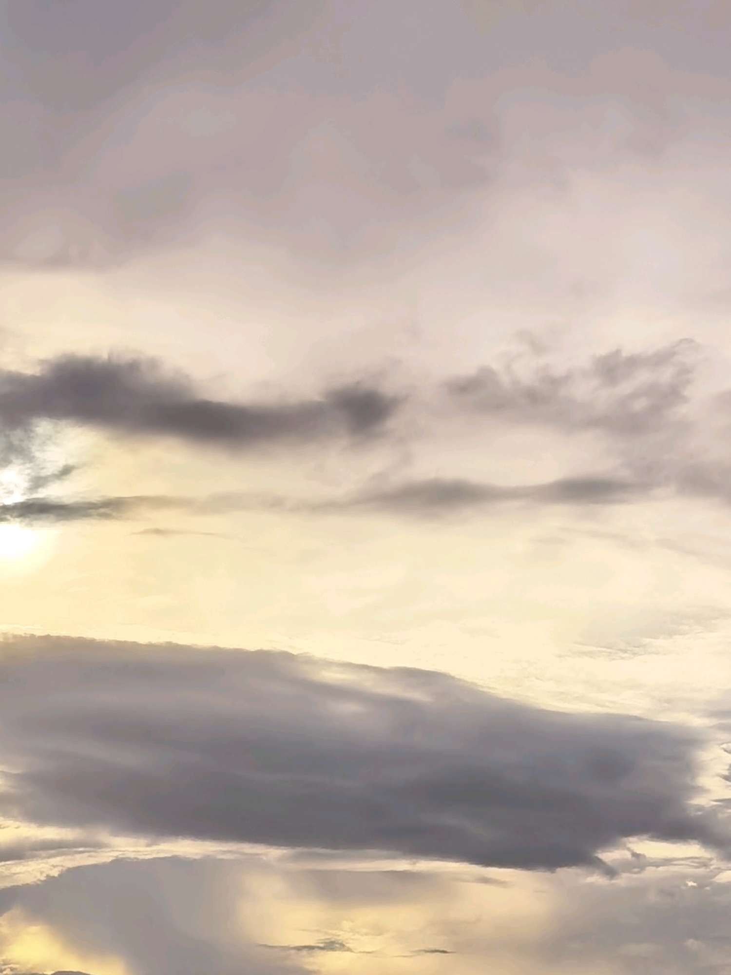 A low-angle, full shot captures a sky filled with wispy clouds at various levels of density. The sun, partially obscured by a thin layer of clouds in the upper left quadrant, casts a soft, yellowish glow across the scene. The clouds range in color from pale gray to a darker, almost purplish hue, suggesting varying altitudes and thicknesses. Some clouds are thin and translucent, allowing the sunlight to pass through, while others are dense and opaque, casting shadows and creating a sense of depth. The overall impression is one of a serene and atmospheric sky, possibly during dawn or dusk, with the light playing a significant role in defining the textures and forms of the clouds. The location is unknown.