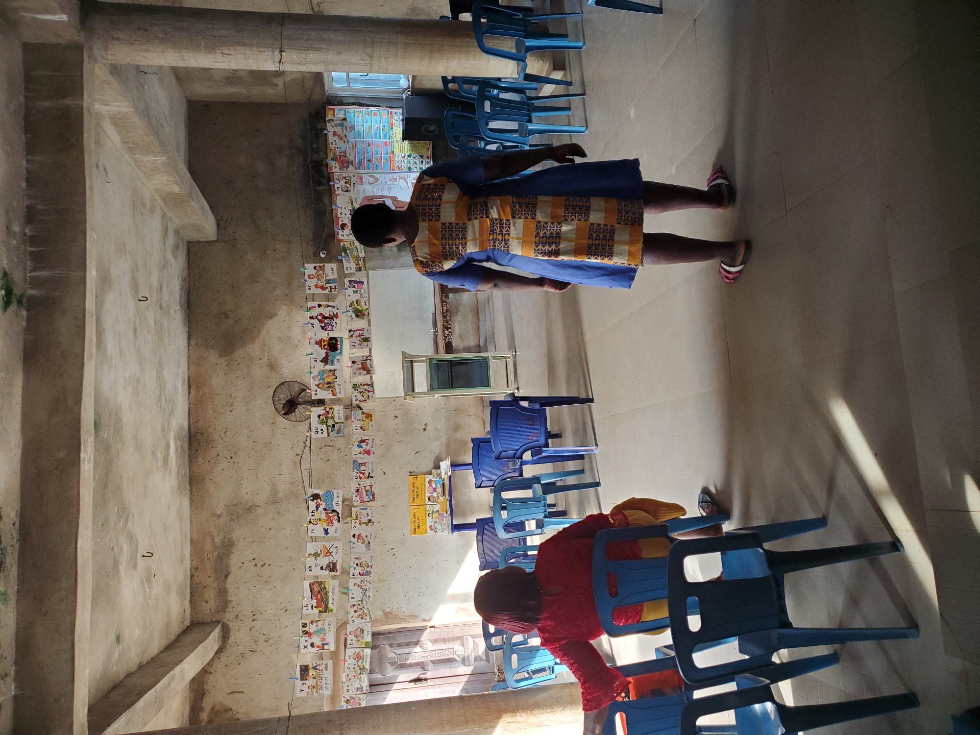 The media file captures an indoor scene, likely a classroom or learning center, in Ukwu-Abwa, Nigeria. The room features concrete walls and floor, with exposed concrete beams in the ceiling, giving it a somewhat utilitarian appearance.
Two individuals are visible. In the foreground, a child, viewed from behind, is seated in a blue plastic chair, wearing a red top. Further back, an adult stands, also viewed from behind, wearing a blue dress with a distinct yellow and dark patterned mid-section, and pink and white sandals.
The room is furnished with numerous blue plastic chairs, some arranged in what appears to be rows, and others stacked. Bright natural light streams in from a window, creating strong sunbeams on the floor.
A prominent feature is a series of educational posters or charts strung along one wall with clothespins. These charts display letters of the alphabet and corresponding words or images, indicative of early childhood education materials. Visible text includes "Y y", "X X N", "ch", "yogurt", "x-ray choo-chao", "H h", "thumb", "QU", "quack", and multiple instances of "M M M M", "K", and "M". Another large chart, possibly an alphabet or vocabulary chart, is partially visible near a window. A small electric fan is mounted on the wall among the educational posters. A few books are stacked on a low shelf or table near the window.