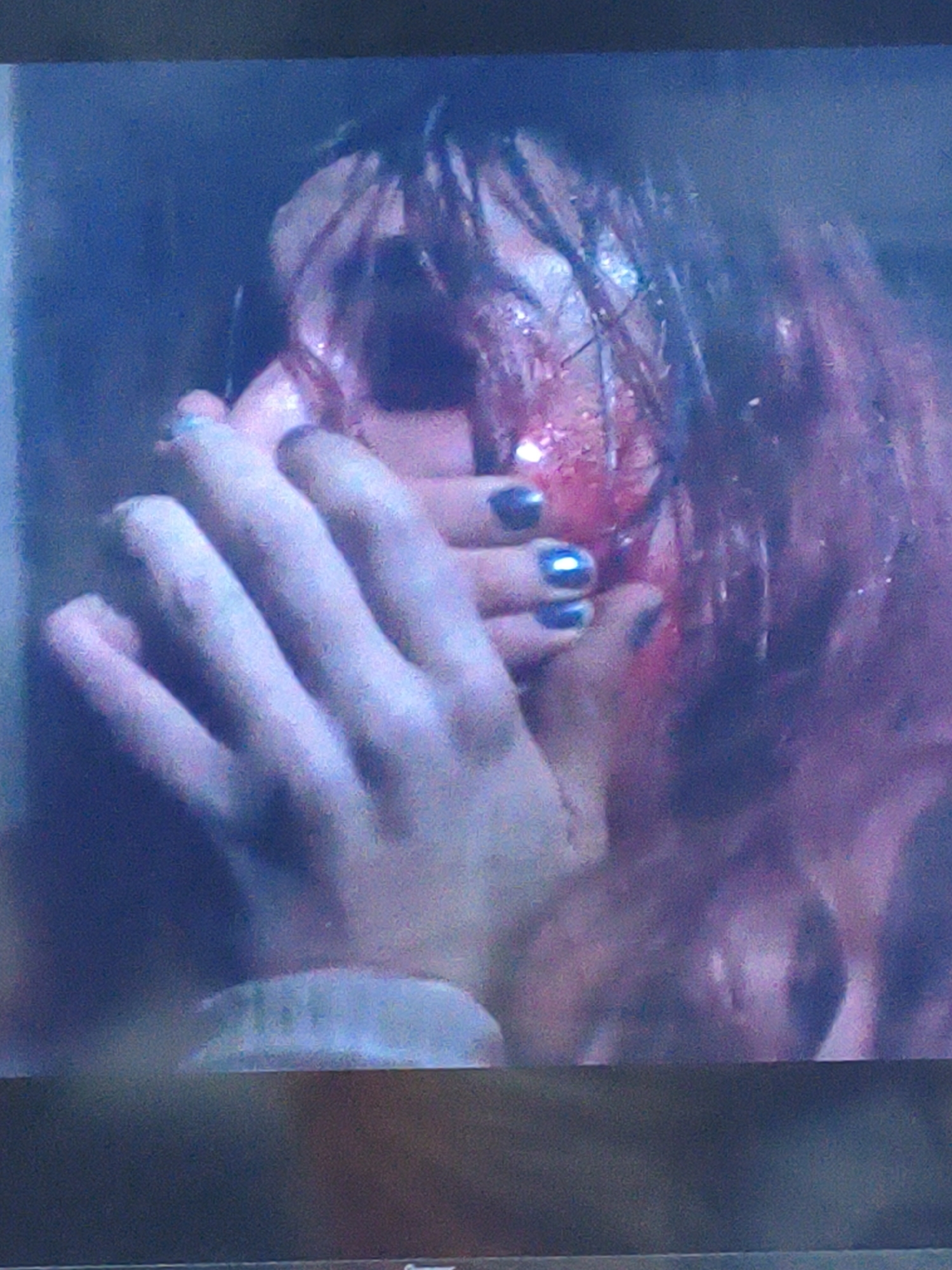 The image shows a close-up, blurred, and low-resolution view of a person's face and hands. The person appears to have dark hair and is wearing dark nail polish on their fingers. The lighting is dim, and the overall scene is dark with blue and purple hues. There are some bright, specular highlights on the person's face and hair, possibly indicating wetness or sweat. The image is heavily pixelated and lacks clear details. No identifiable landmarks of Vladivostok, Russia, are visible in the image.