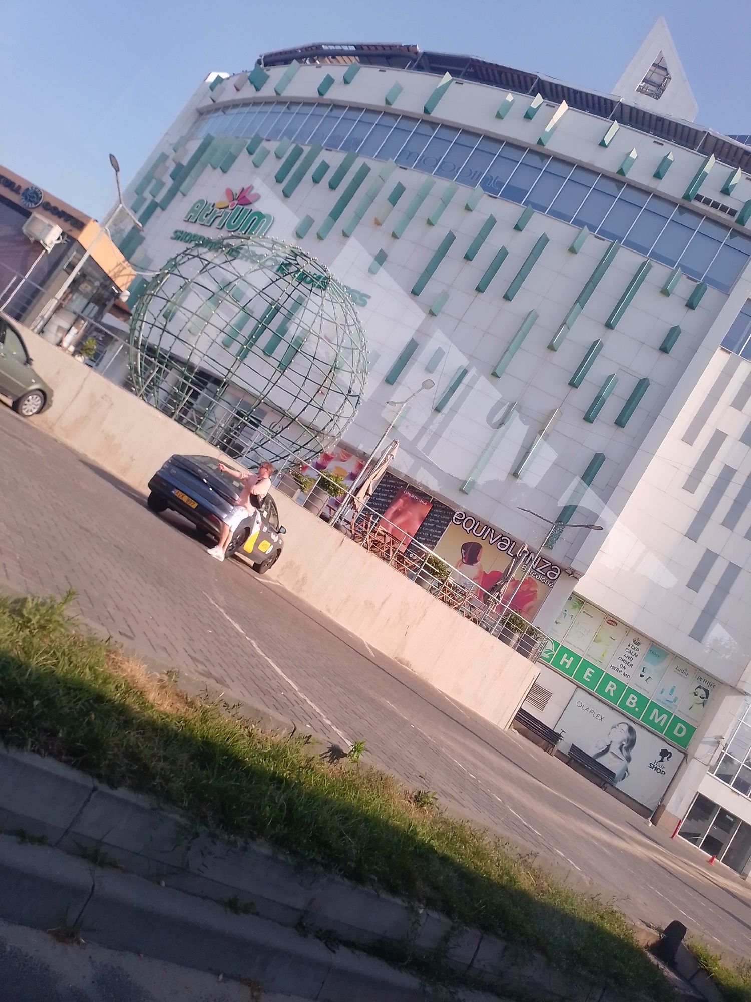 The image shows the "Atrium" shopping center in Chișinău, Moldova, on a sunny day. A modern building with a white facade and green accents dominates the background. In the foreground, a dark-colored car is parked on a cobblestone street. A person, wearing a light-colored top and white shorts, is standing next to the open trunk of the car, seemingly interacting with the contents. A large, green, globe-like sculpture is visible in front of the shopping center. The scene includes a sidewalk with green grass along the edge. Signs for various businesses, including "Equivalenza Barcelona" and "HERB.MD," are visible on the building's facade. The sky is clear and blue.