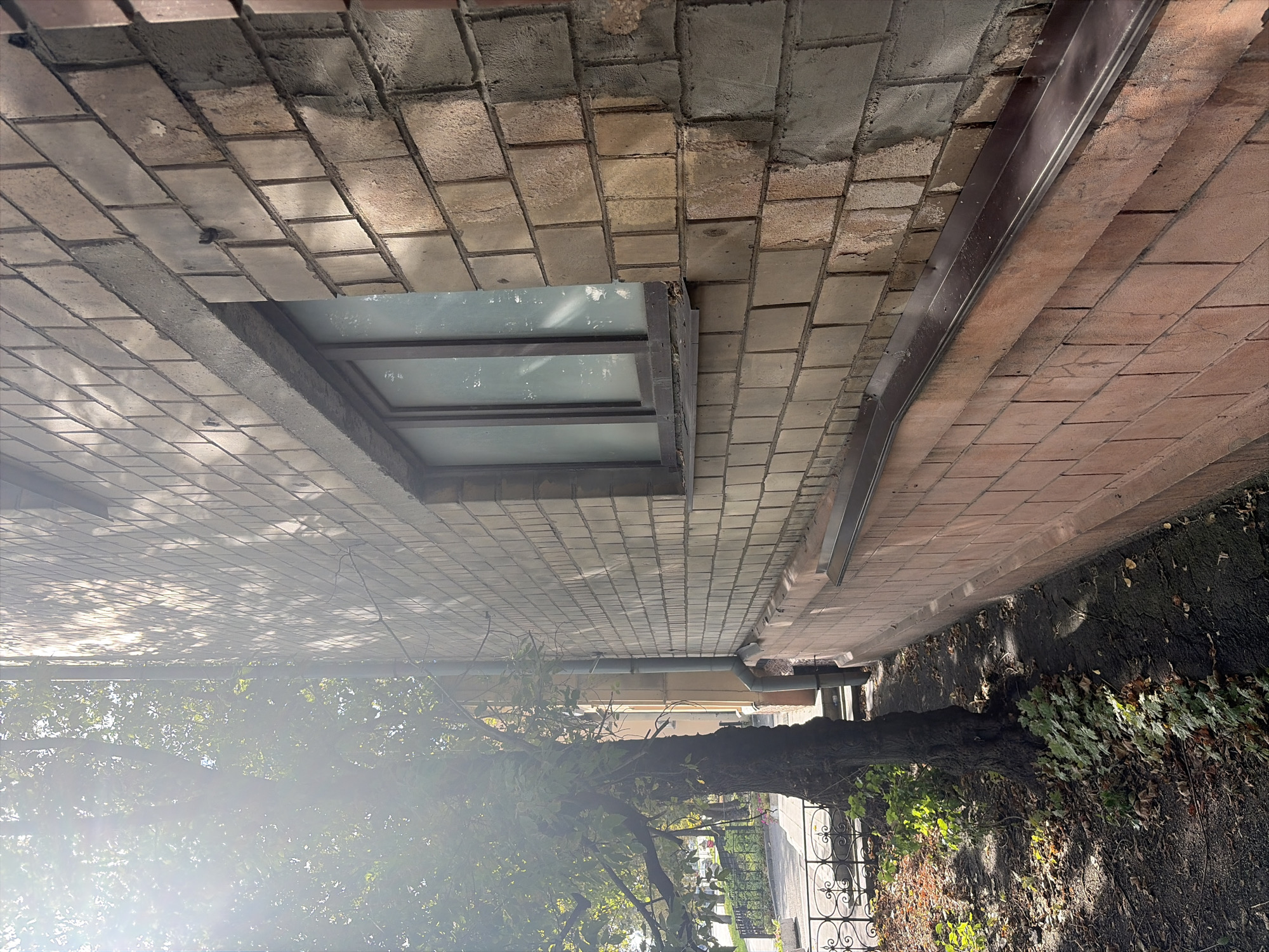 The image depicts a side view of a brick building in Moscow, Russia, with a tree and foliage in the foreground. The building's facade is constructed of beige and pink bricks, with a window featuring frosted glass. Sunlight streams through the leaves of the tree, casting shadows on the brick wall and the ground. The ground appears to be asphalt with fallen leaves scattered on it, suggesting an autumn setting. A metal downspout runs vertically along the side of the building, and a decorative metal fence is visible in the background. The overall impression is one of a quiet urban street corner with natural elements interacting with the man-made environment.