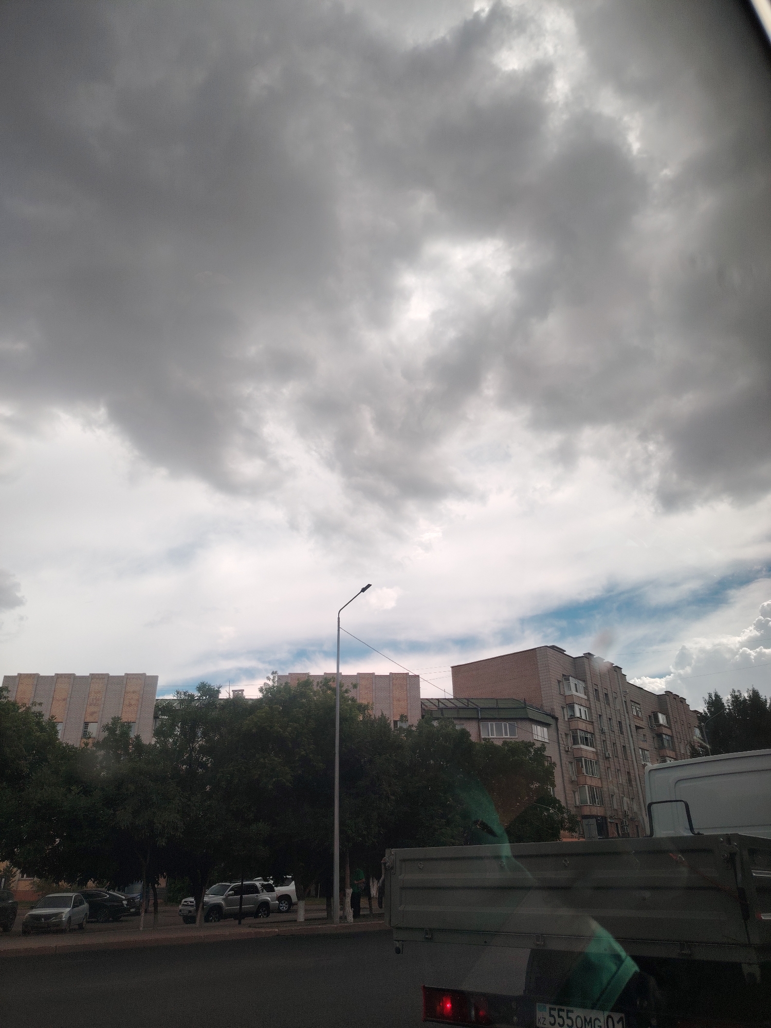 The media file depicts a cloudy sky over a street scene in Astana, Kazakhstan. The sky is filled with dark, heavy clouds, suggesting an impending storm or overcast weather. In the foreground, the back of a grey truck is visible, with part of its license plate showing the text "KZ 555OMG 01". Beyond the truck, several parked cars are visible on the side of the road, including a silver sedan and a white SUV. Trees line the street, and behind them, multi-story apartment buildings are present. One building appears to be under construction or renovation, with its facade featuring a pattern of lighter and darker bricks. The lighting is dim, consistent with a cloudy day. No people are clearly visible in the frame. The overall impression is of an urban environment on a day with challenging weather conditions.