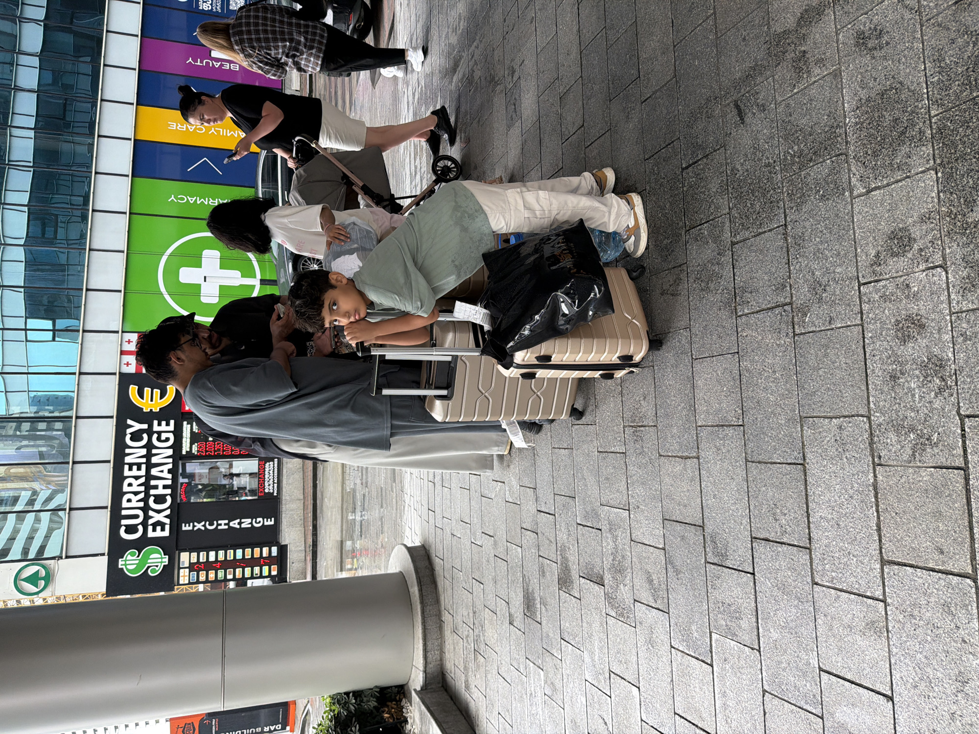 In Batumi, Georgia, a young boy in a green t-shirt and white pants leans against a beige suitcase, looking towards the camera. He is positioned in the foreground, with a black plastic bag also resting on the suitcase. Behind him, a group of people are gathered, some looking at their phones. One woman in a black shirt and white skirt holds her phone, while another woman in a plaid shirt stands further back. A stroller is also visible. In the background, a building with large windows displays signage for "CURRENCY EXCHANGE" and what appears to be a pharmacy or medical clinic with a green cross symbol. The ground is paved with rectangular gray tiles. The scene suggests a public area, possibly near a transportation hub or a commercial district.