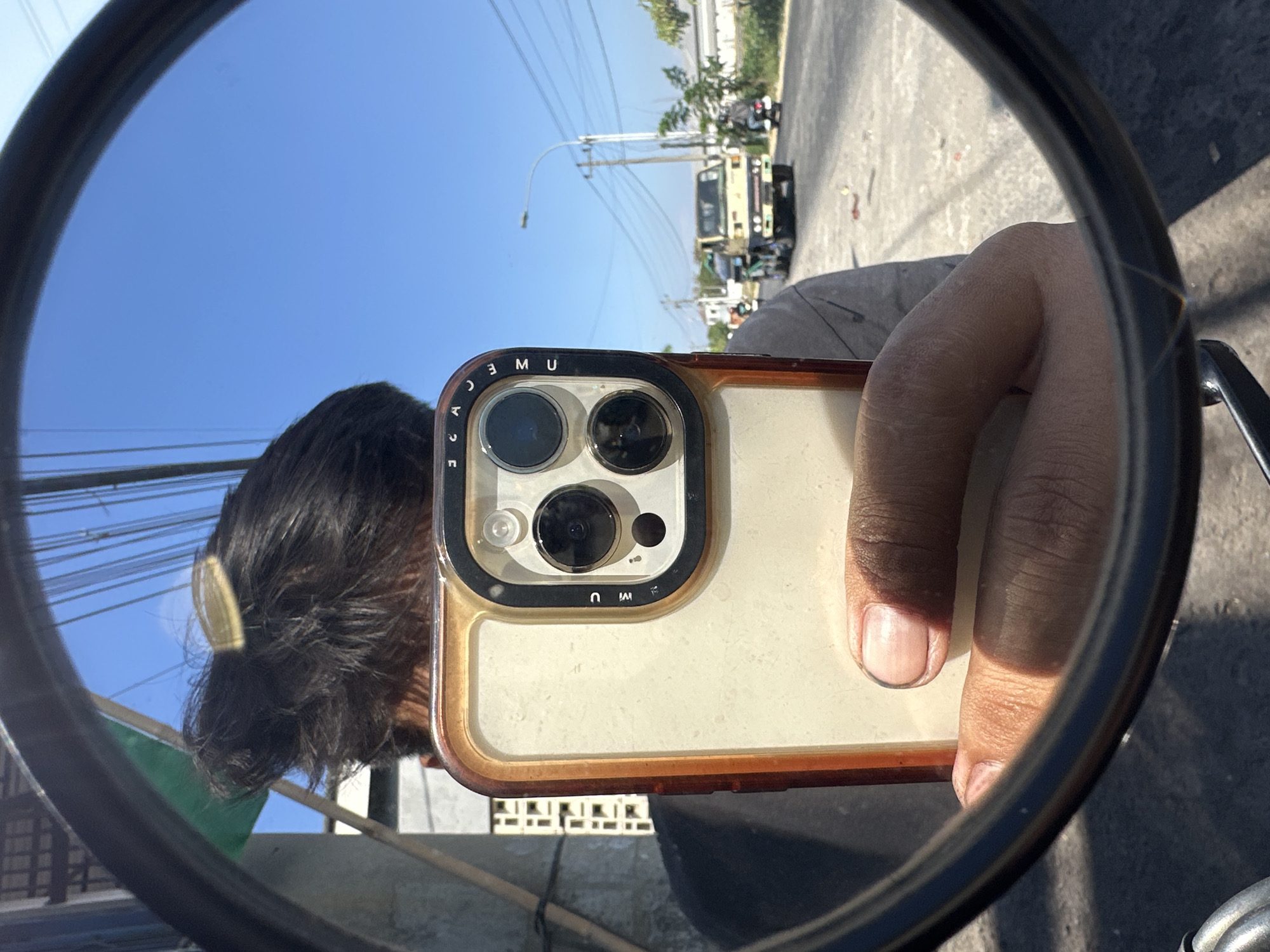 A rear-view mirror reflects a person holding a smartphone, with the camera lens pointed towards the mirror. The person's dark hair is visible, with a gold clip or barrette. The smartphone has a distinctive camera bump with three lenses and a beige case with an orange trim. The reflection shows a street scene with a light-colored truck, two motorcycles, and a lamppost. Buildings and trees line the background, under a clear blue sky. The location is identified as Parangjoro, Indonesia, suggesting this scene is captured on a street within or near that area. The lighting indicates it is daytime, likely sunny.