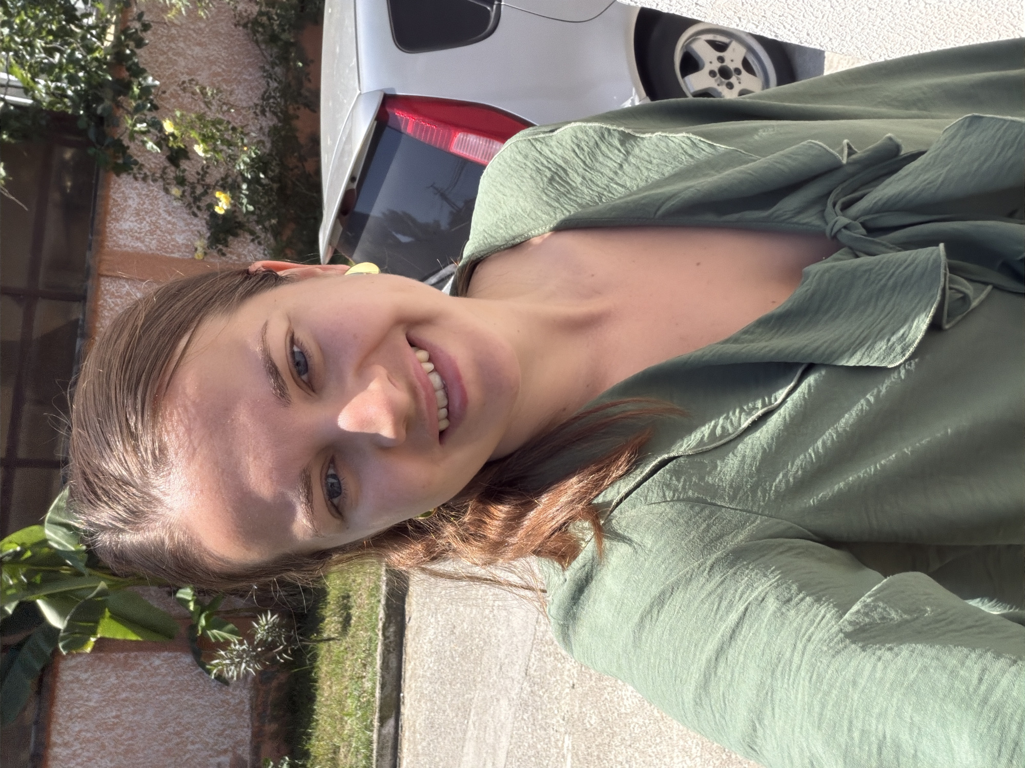 A young woman with brown hair and blue eyes is standing outdoors, possibly in Sochi, Russia, as suggested by the presence of tropical plants. She is wearing a green ruffled shirt and large yellow earrings. The background shows a textured wall, lush green vegetation, and a silver car parked behind her. The lighting indicates it is daytime, with sunlight casting shadows. The woman is smiling and looking directly at the camera, taking a selfie. The overall scene appears to be in a residential or semi-public outdoor area.