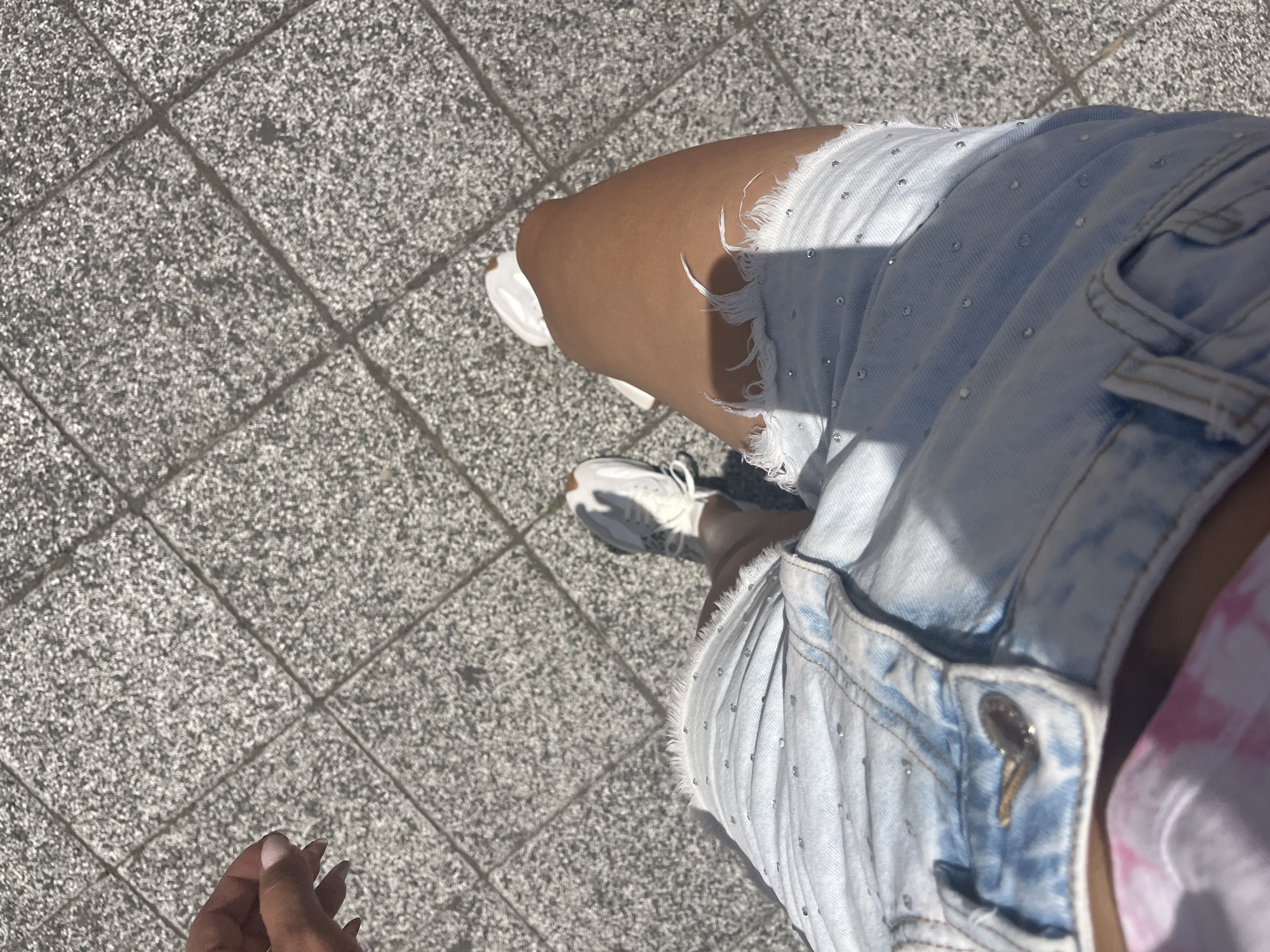 An overhead shot shows a person's legs and hand as they stand on a paved walkway in Plovdiv, Bulgaria. The walkway is composed of square, grey concrete tiles. The person is wearing light blue denim shorts with frayed hems and is adorned with small, scattered rhinestones. White sneakers with visible laces are on their feet. A hand with manicured nails is reaching out from the left side of the frame, close to the ground. Shadows cast by the sun are visible across the tiled surface. The lighting suggests it is daytime.