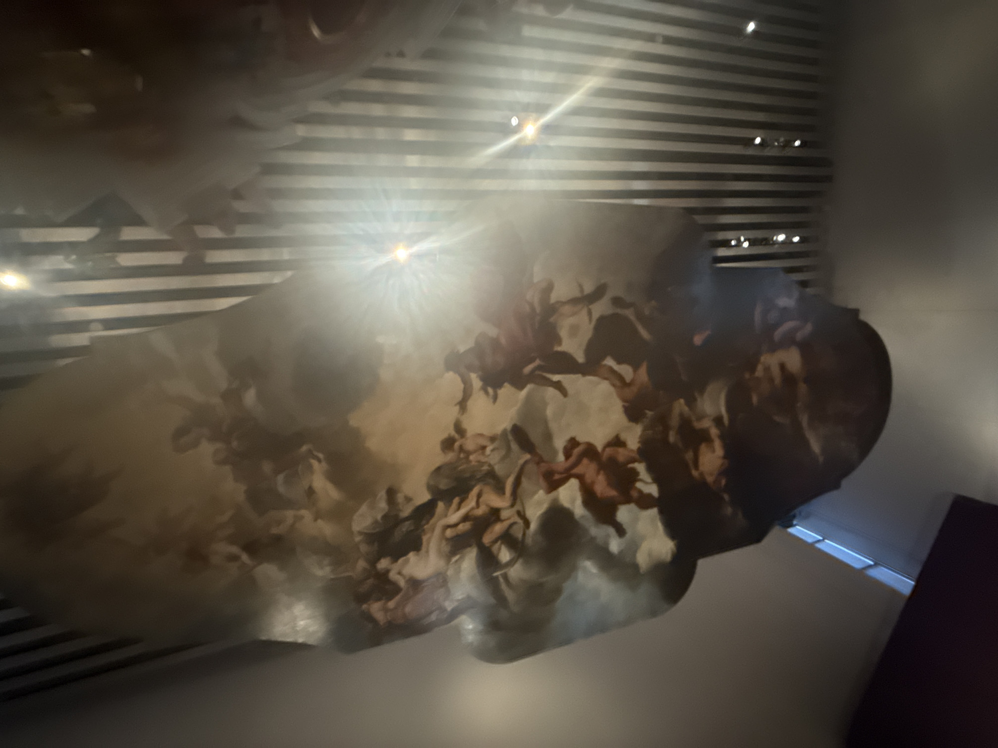 The image shows a ceiling with a painted mural in San Bernardino Tlaxcalancingo, Mexico. The mural depicts numerous human figures, possibly angels, in a cloud-like formation. The ceiling also has a modern striped design above the mural. A light source, likely a spotlight, shines directly onto the mural, creating a bright glare. Part of a dark-colored wall is visible in the lower right corner.