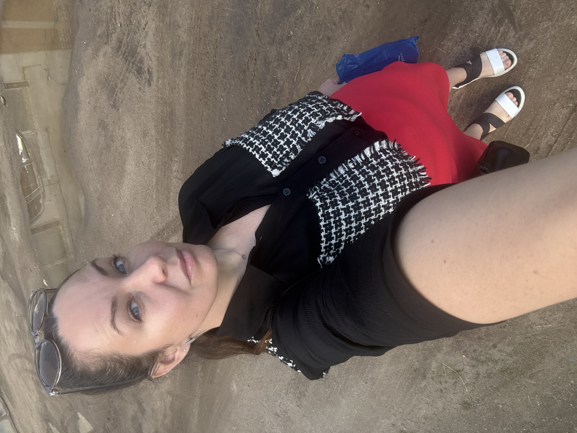 A woman is taking a selfie outdoors, holding the camera with her right arm. She is wearing a black and white tweed jacket over a black top, a red skirt, and white and black sandals. She has sunglasses perched on top of her head and is wearing a delicate necklace. Her hair is pulled back in a ponytail. She is standing on a dirt or gravel path, and in the background, there is a large puddle of water that reflects an industrial-looking building with windows. This scene is consistent with the industrial areas or waterfront along the Kama River in Chaikovsky, Russia, which is known for its oil and chemical industries and has associated infrastructure and water bodies.