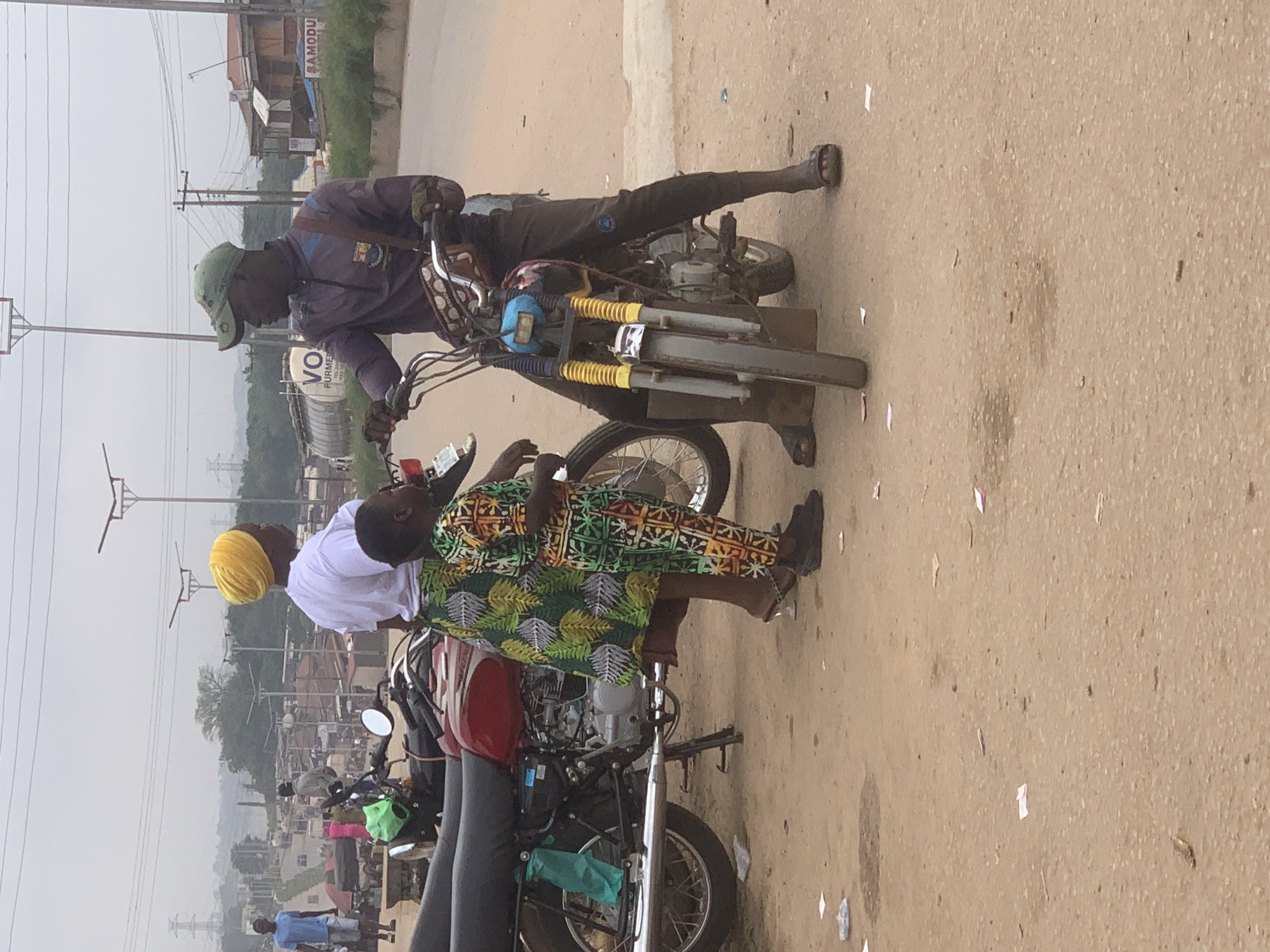 In Shagamu, Nigeria, on a cloudy day, three individuals are gathered around two motorcycles on a dusty, unpaved street. The foreground is dominated by the motorcycles and the people. To the left, a red motorcycle is parked, its rear section visible. A woman wearing a white top and a vibrant green and yellow patterned skirt stands next to it. She has a yellow headscarf. To her right, a young child, dressed in a colorful, patterned jumpsuit, stands and interacts with the woman.
To the right of the scene, a man wearing a dark purple jacket, dark pants, and a green cap is seated on a motorcycle. The motorcycle has a blue front light and yellow accents on its front suspension. He is facing to his left, seemingly in conversation with the woman. The man has a patterned bag slung across his chest.
The background reveals a bustling environment. Beyond the motorcycles, there are more motorcycles, tricycles, and various market stalls or shops. Buildings line the street, and some industrial structures, including large cylindrical tanks, are visible further in the distance. Power lines and streetlights are also present in the background, indicating an urban or semi-urban setting. The ground is covered in dirt and scattered with small pieces of paper or debris. The overall impression is of a typical street scene in a Nigerian city, possibly a commercial or transportation hub.