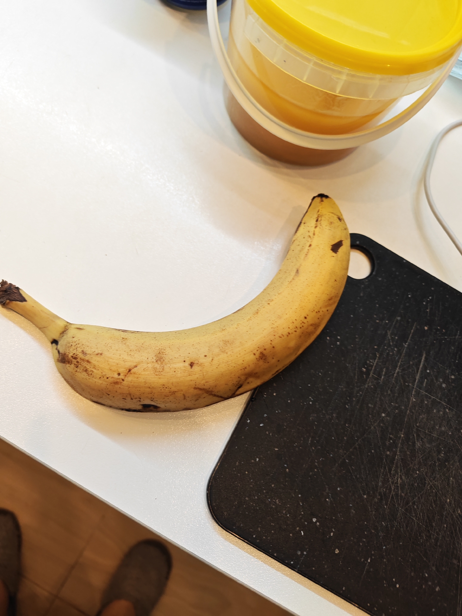 A slightly bruised, yellow banana rests on a white countertop, adjacent to a black cutting board. To the upper right, a yellow-lidded plastic container, likely holding a food product, is visible. A white electrical cord is also present in the background. The scene is illuminated by overhead lighting, casting a soft glow. The overall impression is of a simple, domestic setting, possibly a kitchen. There is no indication of the location being in Bataysk, Russia, nor are there any individuals present in the image.