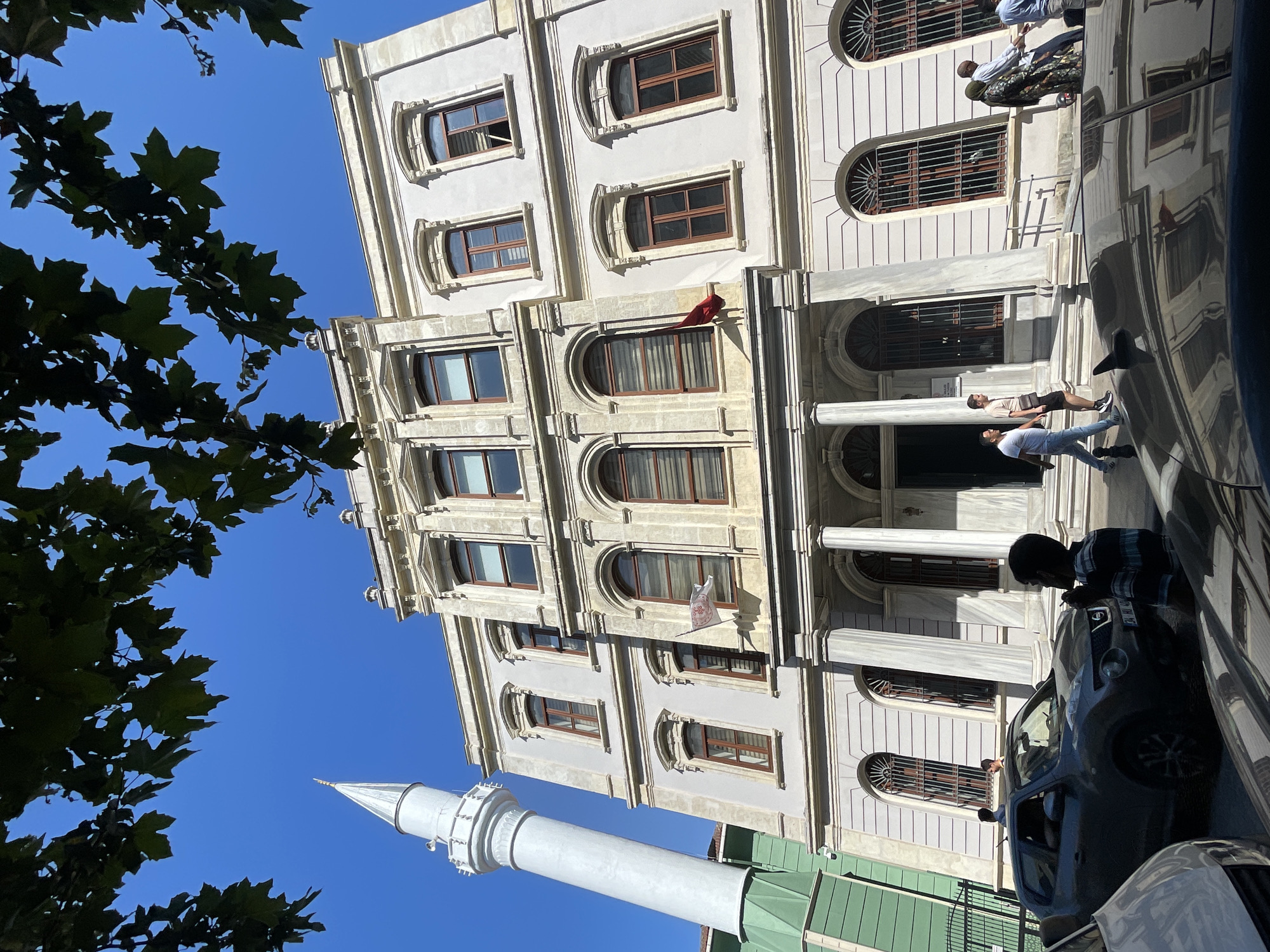 The image captures a view of a historical building with a minaret in Istanbul, Turkey, on a bright, sunny day. The building is a light-colored, multi-story structure with classical architectural elements, including columns and arched windows. A tall, slender white minaret stands to the left of the building. In the foreground, several cars are parked on a street, and a few people are visible walking or standing near the entrance of the building. One person, a young male wearing a striped shirt, is standing near a dark-colored SUV, appearing to smoke. Another group of people, including a woman in a patterned dress and headscarf, are walking past the building entrance. The sky is clear and blue, and the upper portion of the image is partially obscured by dark green tree leaves, suggesting the presence of trees in the vicinity. The overall scene suggests a public or administrative building in an urban setting within Istanbul.