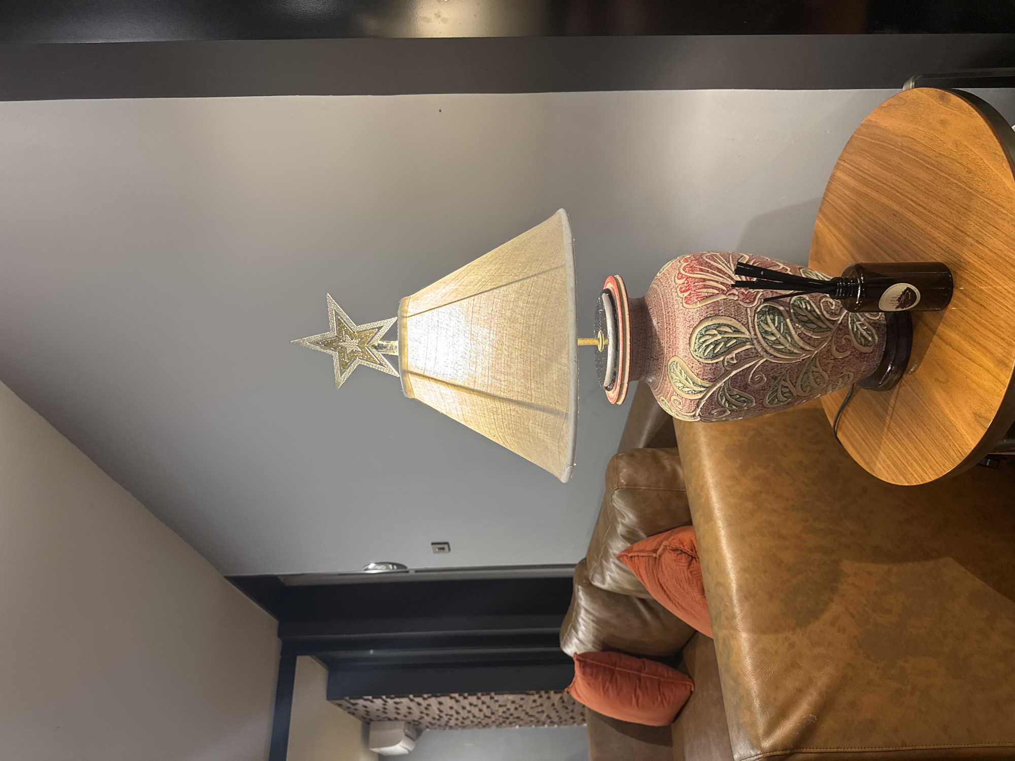 In a contemporary living room setting, likely in Bangkok, Thailand, a warmly lit table lamp with a star ornament sits on a small, round wooden table. The lamp's shade is a neutral beige, and its base is a decorative ceramic vase featuring a floral motif in shades of pink, green, and red. Next to the lamp on the table is a small dark glass bottle with diffuser reeds, suggesting a home fragrance. Behind the table, a plush, brown leather sofa with two orange accent pillows is visible. The room's walls are painted in a cool grey, and a black vertical column is present on the right side of the frame. In the background, another part of the room shows an air conditioning unit mounted on a wall with a textured, mosaic-like pattern. No people are present in the scene.