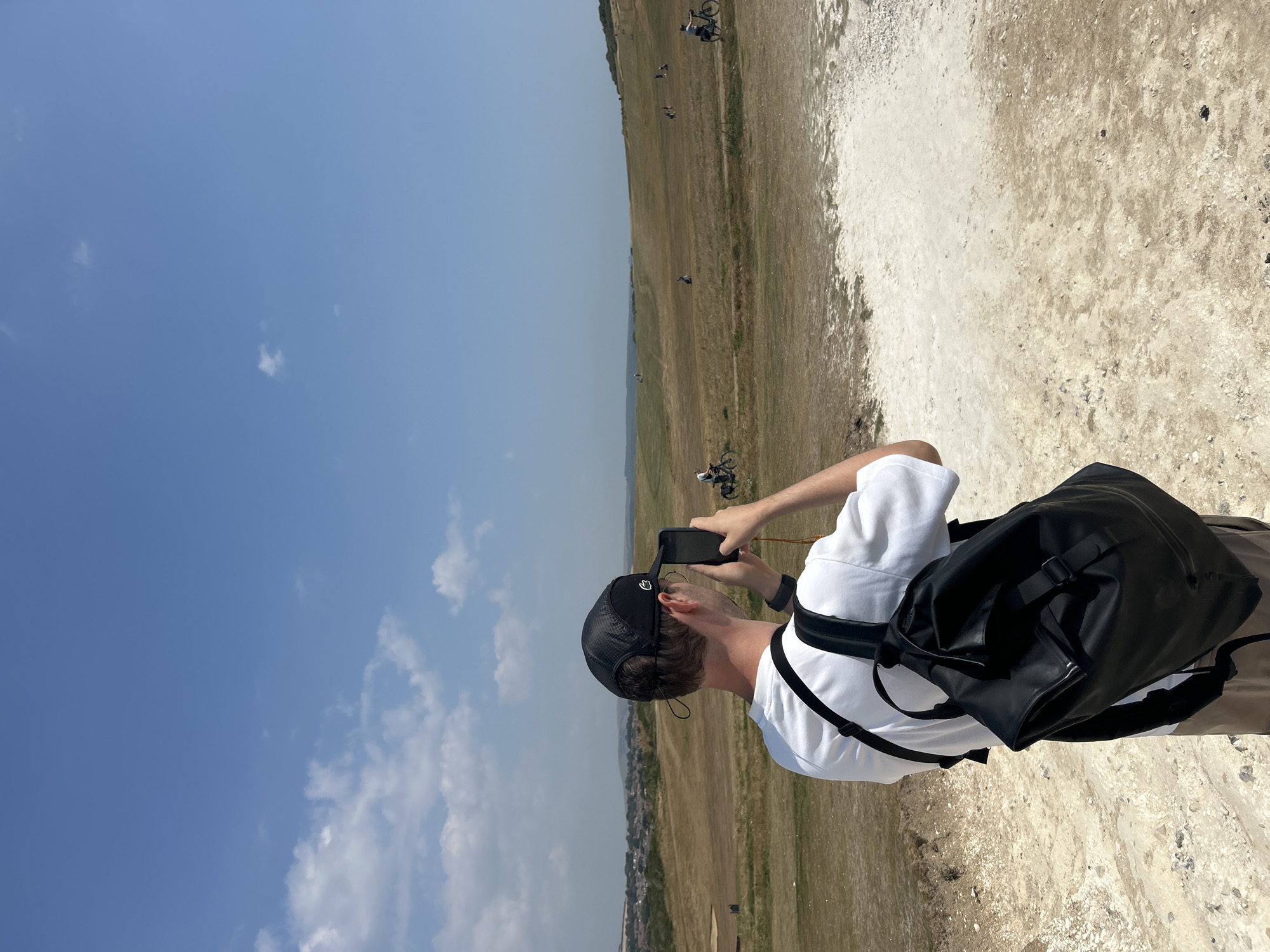 A man wearing a white t-shirt and a black baseball cap is standing and taking a photo with his phone in a landscape with dry grass under a clear blue sky with some clouds. He has a black backpack on. In the background, there are several people on bicycles and a town in the distance. The foreground consists of dry, sandy ground with some rocks.