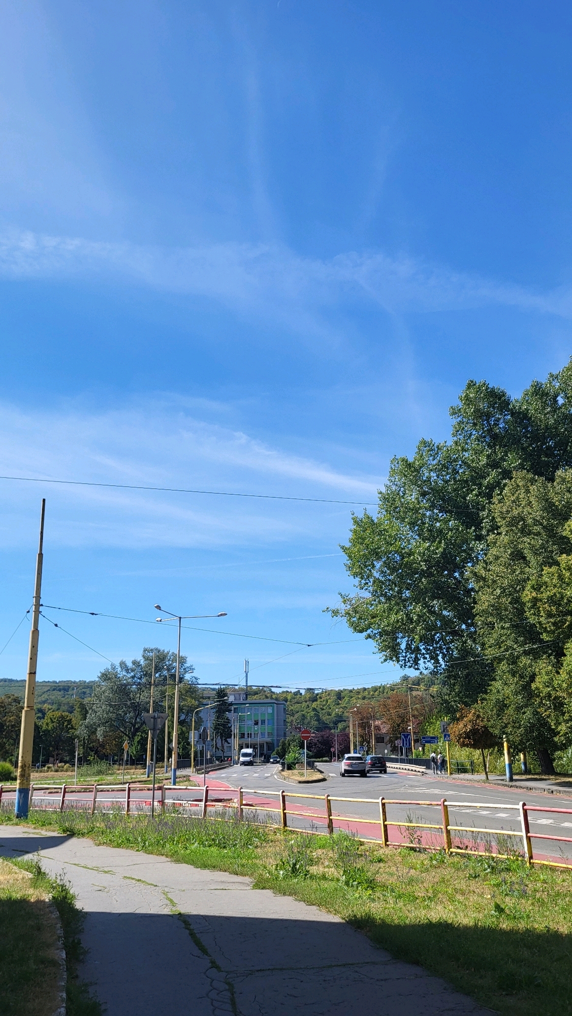 The image depicts a street scene in Prešov, Slovakia, on a clear day with a bright blue sky. A paved pathway is in the foreground, leading towards a road. The road has multiple lanes and is lined with a red and yellow guardrail. Cars are visible on the road, including a white van, a silver car, and a dark car. Pedestrians are also present, walking on the sidewalk.
On the left side of the road, there are utility poles and trees. A building, possibly a school or office, with green accents, is visible in the background. Trees and vegetation are abundant throughout the scene, with a large, leafy tree dominating the right side of the image. The overall impression is a typical urban environment with elements of nature interspersed. The location is likely in a residential or commercial area of Prešov, given the presence of a building and the road infrastructure.