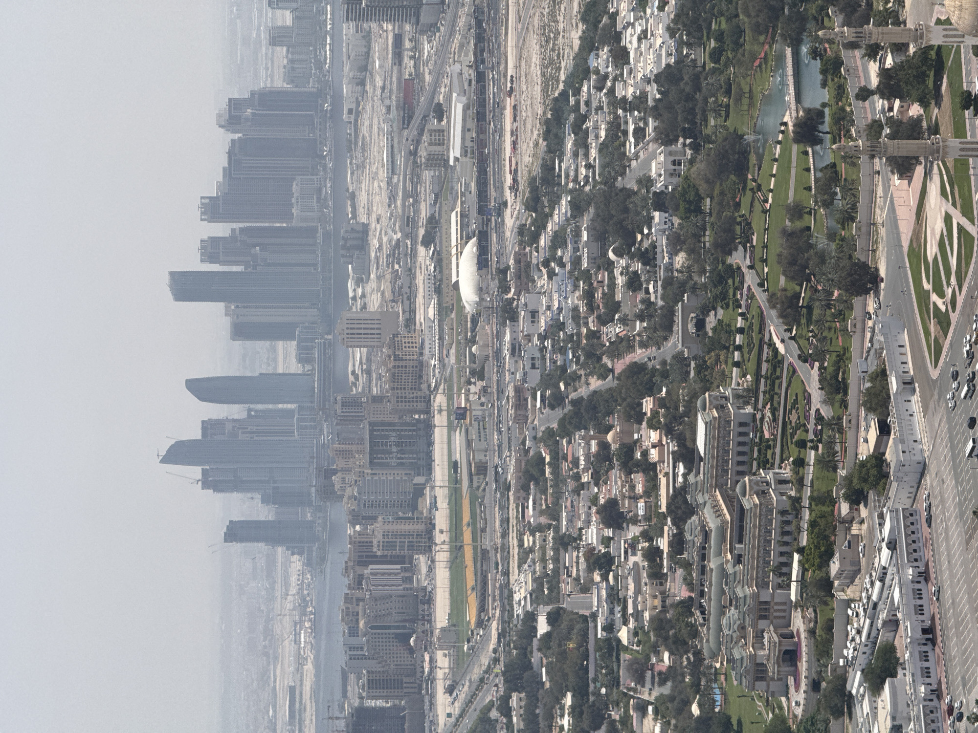 This aerial photograph captures a vast and dynamic urban landscape identified as Dubai, United Arab Emirates. The image is taken from a high vantage point, looking down over an expansive city that seamlessly blends modern skyscrapers, extensive residential areas, and manicured green spaces.
The left side of the frame is characterized by a dense concentration of tall, contemporary skyscrapers, many of which appear to be under construction, with visible cranes hinting at ongoing development. These high-rise buildings gradually fade into a prominent atmospheric haze, rendering the most distant structures faint and indistinct.
A wide, multi-lane highway system traverses the middle section of the city, bustling with numerous vehicles. Along this central corridor, a mix of commercial and residential structures is visible, alongside a large, vibrant green sports stadium or field. Several prominent white buildings with distinctive architectural features punctuate the urban fabric. Notably, a white billboard with red text displaying "Dower.ru" can be seen near the highway, and the word "EMAAR" is discernible on a long, white building.
The right side of the image unfolds into a more verdant scene. A large, intricately designed green park dominates this section, featuring winding pathways, lush trees, and a serpentine water body with visible fountains. Adjacent to the park, several elegant minarets, indicative of mosques, stand tall. Beyond the park, extensive residential districts comprising numerous smaller, uniform buildings, likely villas or townhouses, are interspersed with greenery.
The overall impression is that of a rapidly expanding and well-planned modern city. The prevailing atmospheric conditions appear hazy or dusty, reducing clarity in the distance. The lighting suggests it is daytime, possibly late morning or early afternoon, with minimal harsh shadows.