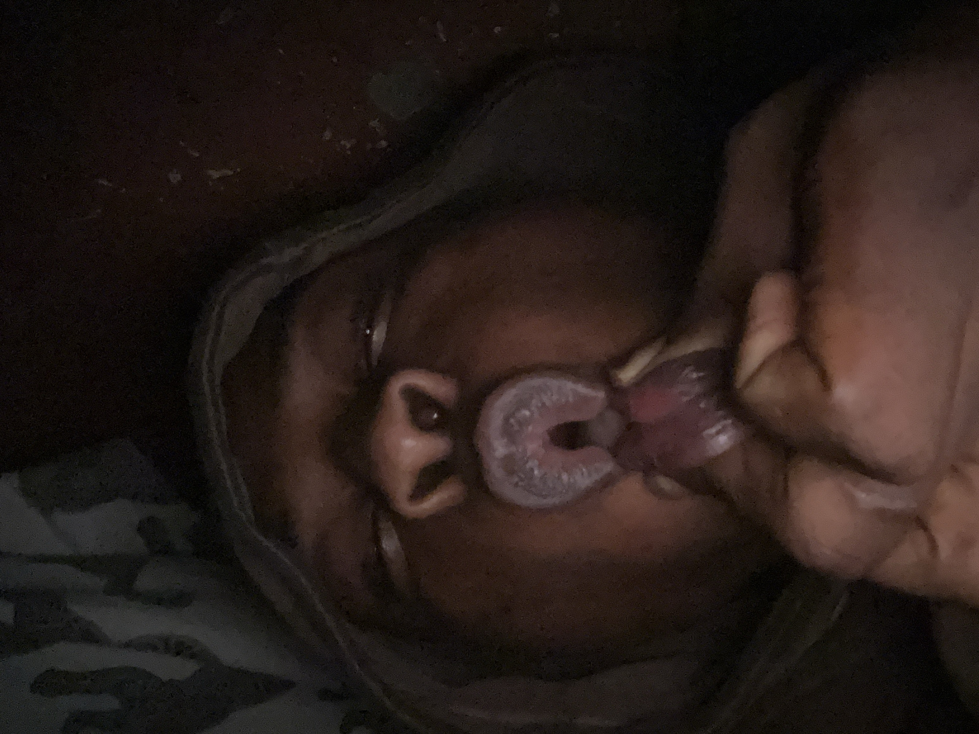 A close-up, low-light, overhead shot captures a person with dark skin in Ikorodu, Nigeria. Their eyes are closed, and their mouth is open, with a tongue visible inside. A finger is touching the tongue. The background is dark and out of focus, with some indistinct patterns that might be fabric.