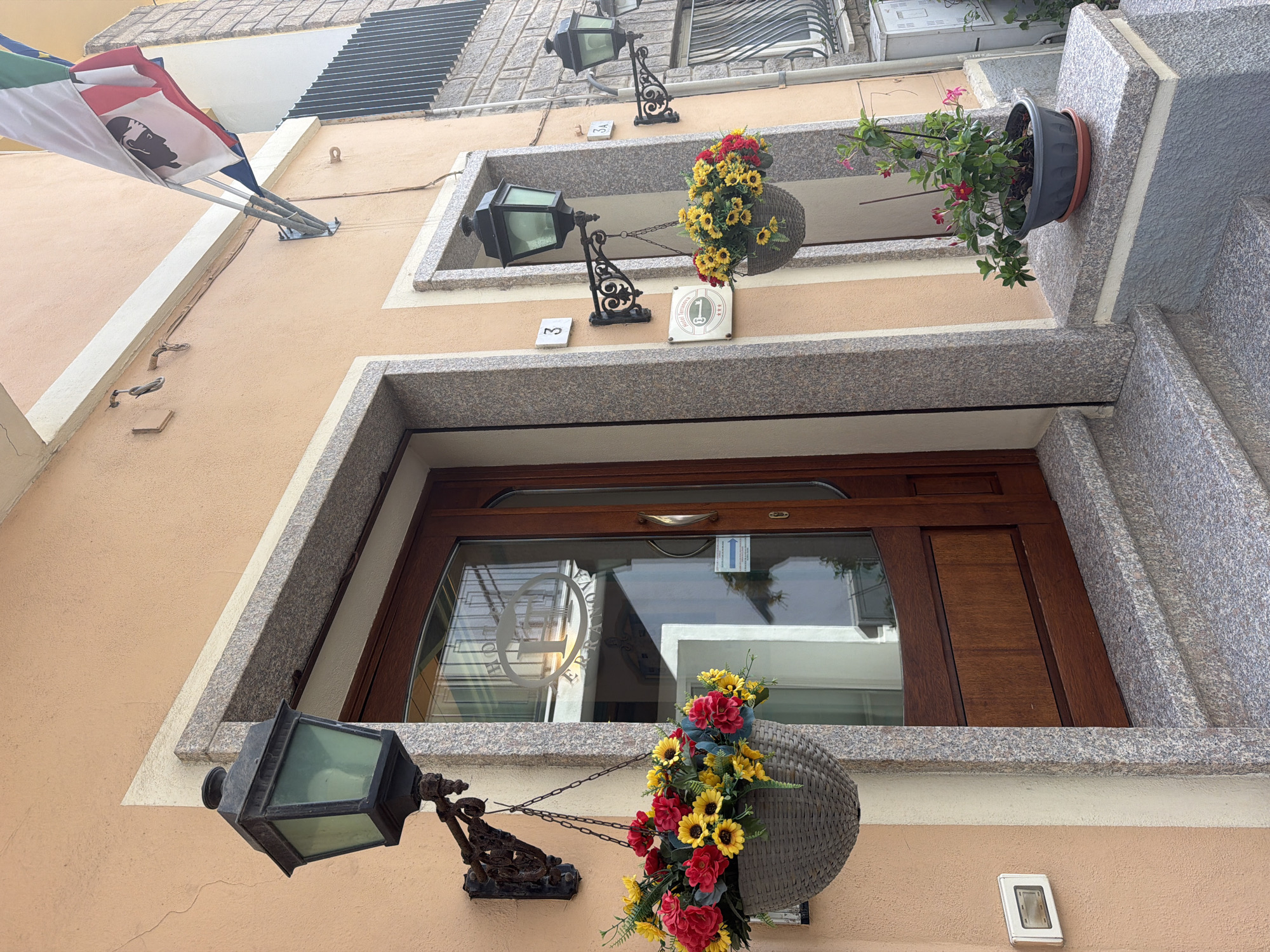 The image depicts the exterior of a building, likely a hotel given the signage, located in Olbia, Italy. The facade is painted in a warm peachy hue, with a prominent wooden door framed by granite. Two black, traditional style lanterns hang on either side of the entrance, each adorned with a hanging basket filled with vibrant red and yellow flowers. Above the door, a flag flies, featuring a white background with red and green stripes and a profile of a black head. To the right of the main entrance, a smaller, similar doorway is visible, also featuring a lantern and a hanging flower basket. A potted plant with pink flowers sits on a granite ledge to the right of the smaller entrance. The building's architecture suggests it is situated in a Mediterranean coastal town. The scene is bright, indicating daylight.
