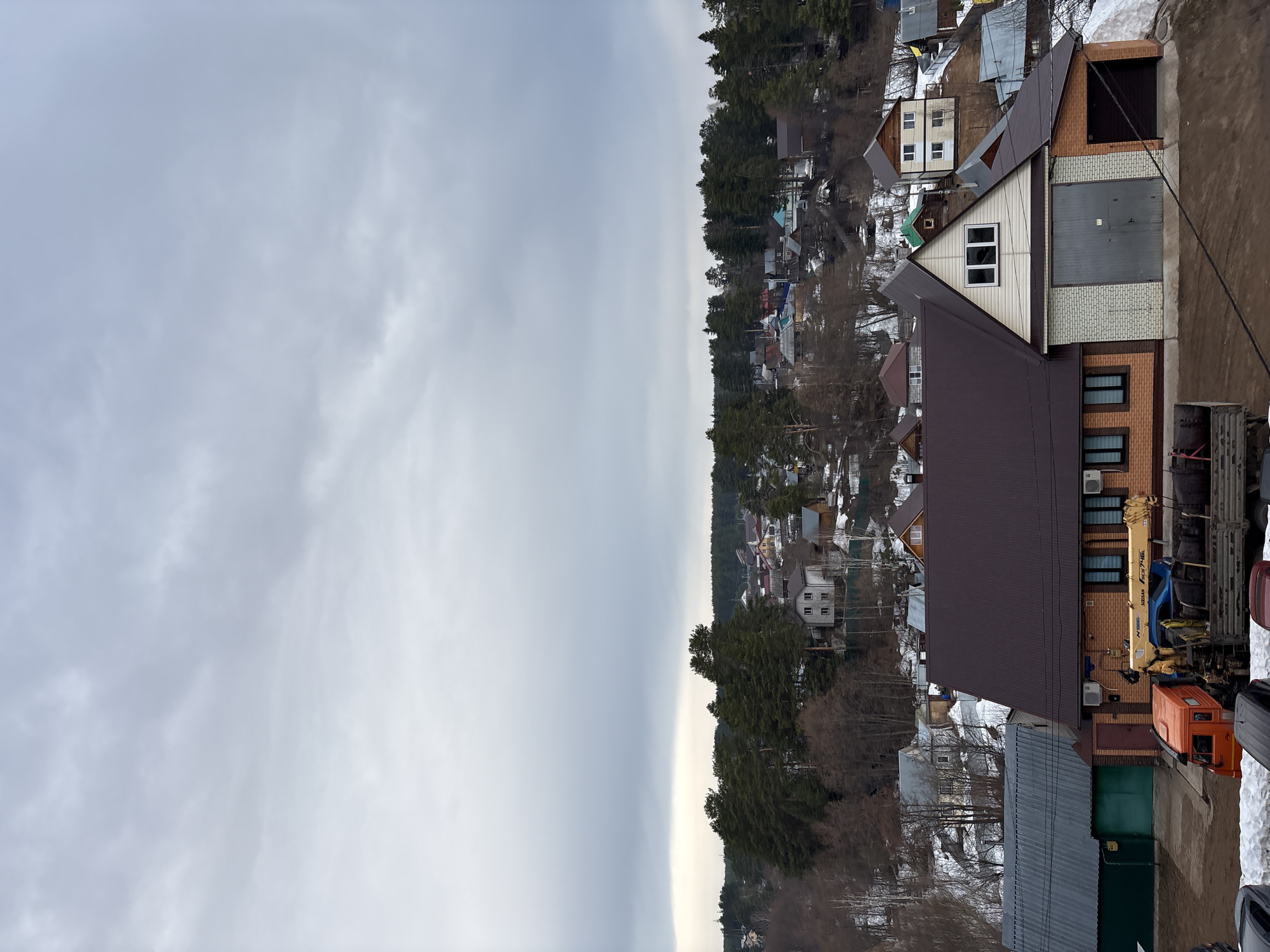 The image shows a view in Chaikovsky, Russia, likely from an elevated position, perhaps a building or hill. The sky is overcast, with grey clouds filling the left side of the frame. On the right side of the image is a residential area. Houses are visible, interspersed among trees. The housing appears mixed in style and size, some with snow-covered roofs, suggesting it may be a somewhat rural part of the town. Some trees are bare, indicating a possible late winter or early spring season. In the foreground, the roofs of buildings are visible, along with a crane truck with an orange cab.