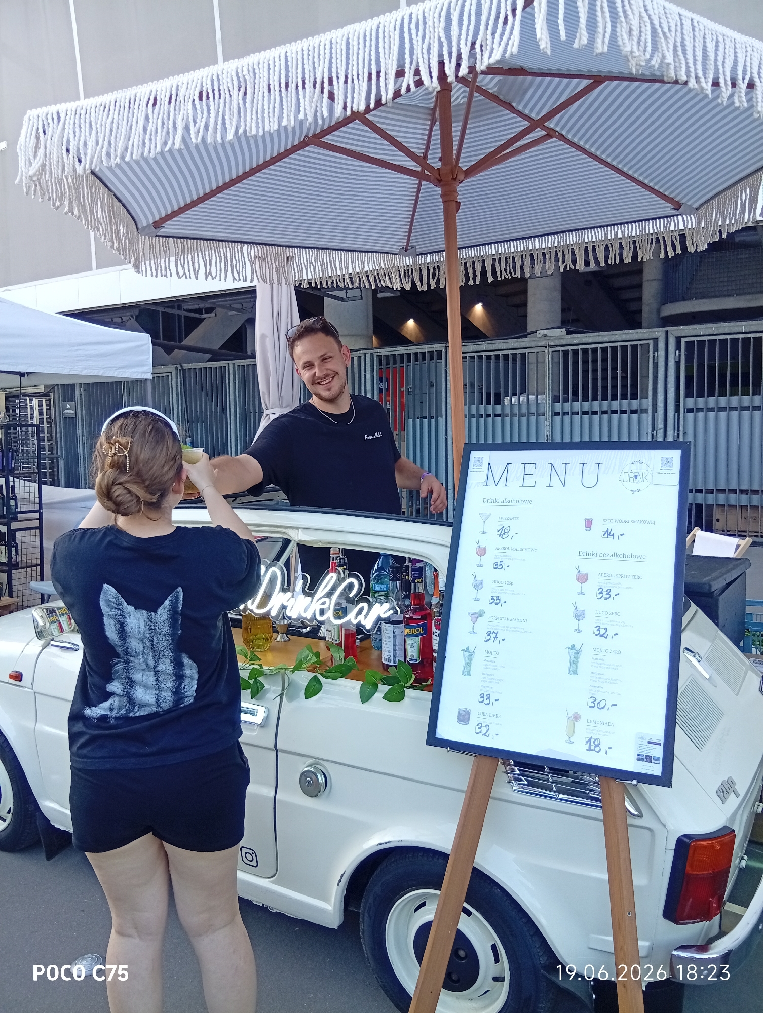 A man and a woman are standing by a white car converted into a bar. The car has a striped umbrella over it. The man is handing a drink to the woman. A menu board stands in front of the car. The scene appears to be outdoors, with a fence and stadium seating in the background. The text on the menu and the car indicate the location is "DrinkCar" and "Mojito." The date "19.06.2026" and time "18:23" are visible at the bottom right. The text on the woman's shirt is a black and white image of a cat.