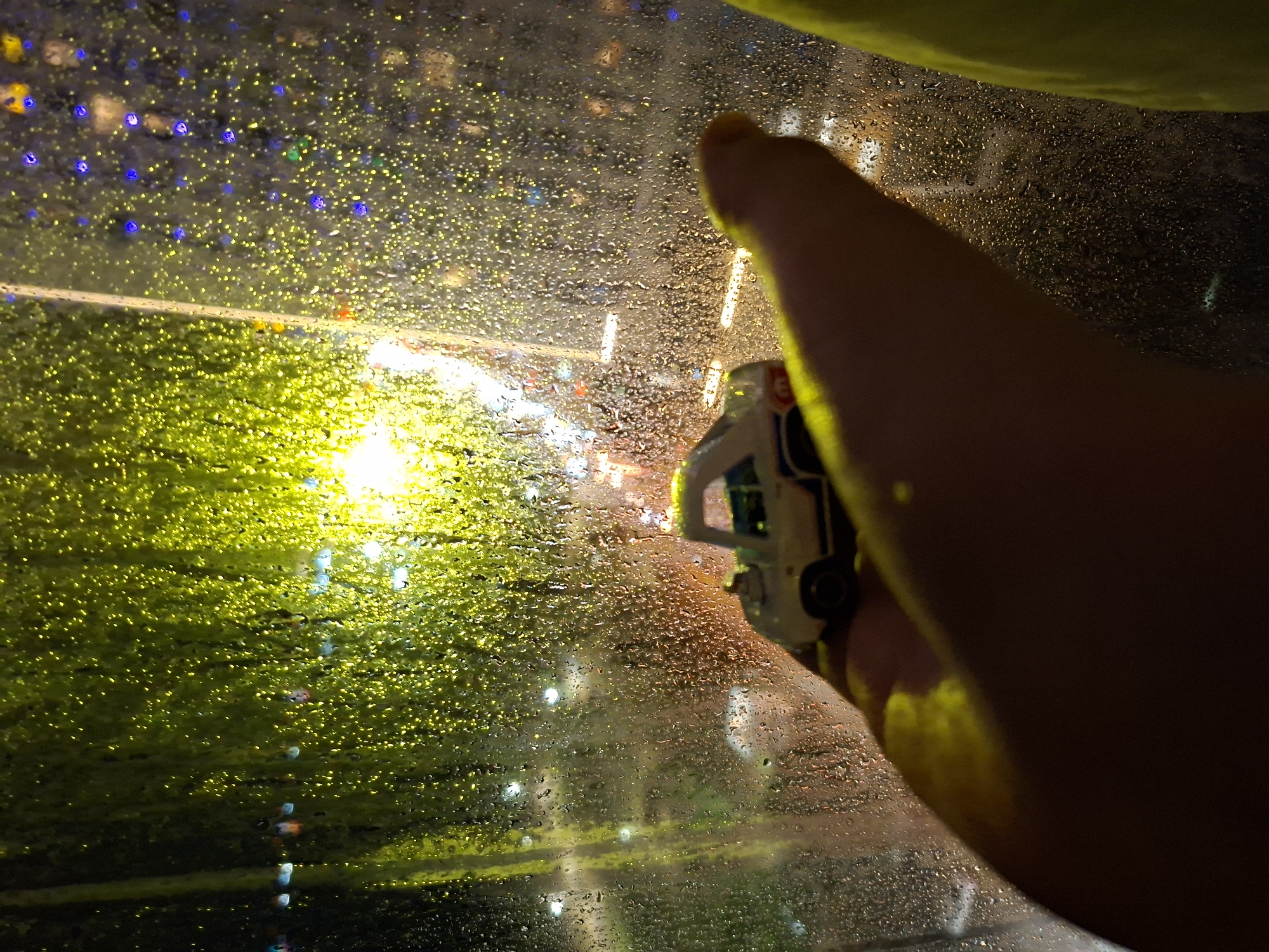 A person's hand holds a miniature white and blue car against a rain-covered window at night. Outside the window, city lights are visible, including a yellow illuminated area, suggesting a street or road. Bokeh-like effects obscure the background details. The location is inferred to be Nha Trang Ward, Vietnam, based on image context.