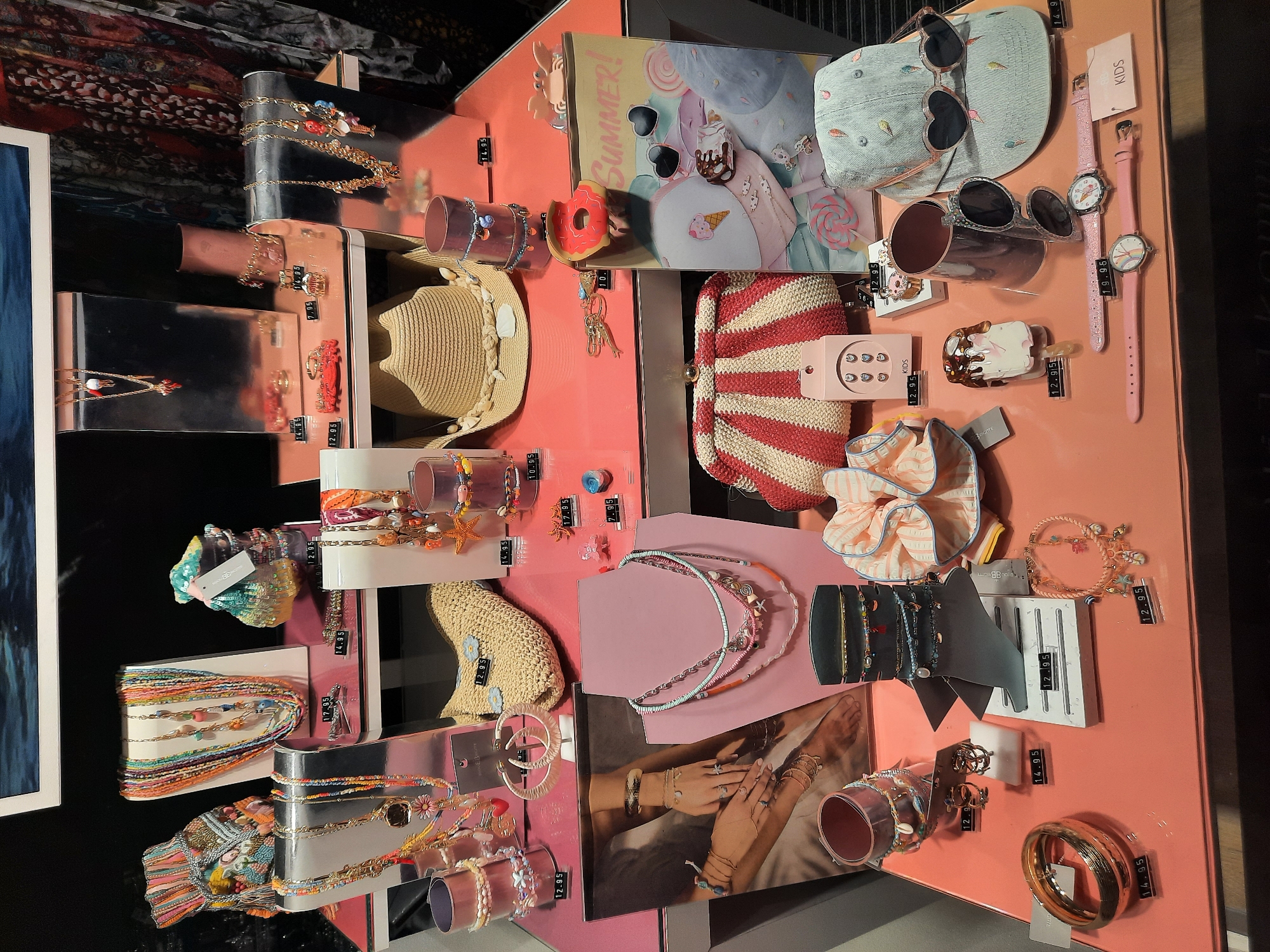 This is a retail display of summer-themed accessories, likely in a store in Rosenheim, Germany. The display is set up on a coral-colored surface and features a variety of items including necklaces, bracelets, a straw hat, a clutch bag, sunglasses, watches, and small decorative items like a donut charm and a crab figurine. Many of the accessories have a nautical or beachy aesthetic, with charms shaped like shells, starfish, and ladybugs. Prices are displayed on small black and white tags in front of some of the items. The lighting is bright and highlights the colorful merchandise. No people are visible in the image.