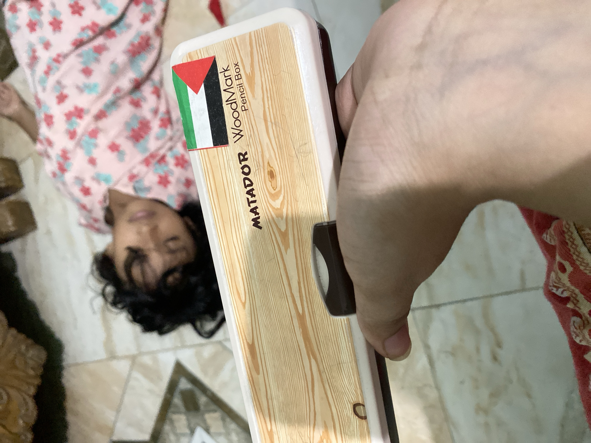 A person, whose gender and age are not clearly discernible due to poor image quality and a closed eye, lies supine on a tiled floor. They are wearing a pink and blue floral-patterned garment. In the foreground, a hand holds a white pencil box with a wood-grain design. The pencil box features the "MATADOR WoodMark Pencil Box" branding and a Palestinian flag sticker. The environment appears to be indoors, with a partially visible ornate furniture piece in the background. There is no clear indication of the specific location within Dhaka, Bangladesh.