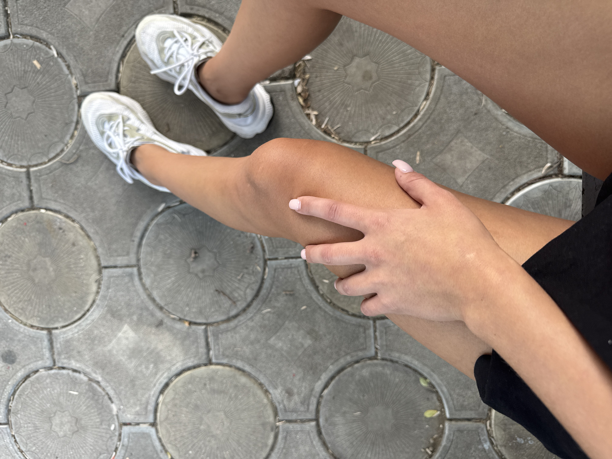 A top-down view captures a person's legs and feet wearing white sneakers, resting on a patterned gray pavement. A hand with lightly colored nail polish is placed on the left thigh. The pavement consists of interlocking square tiles, each featuring a circular relief pattern with radiating lines. The scene is outdoors, with some debris visible on the ground.