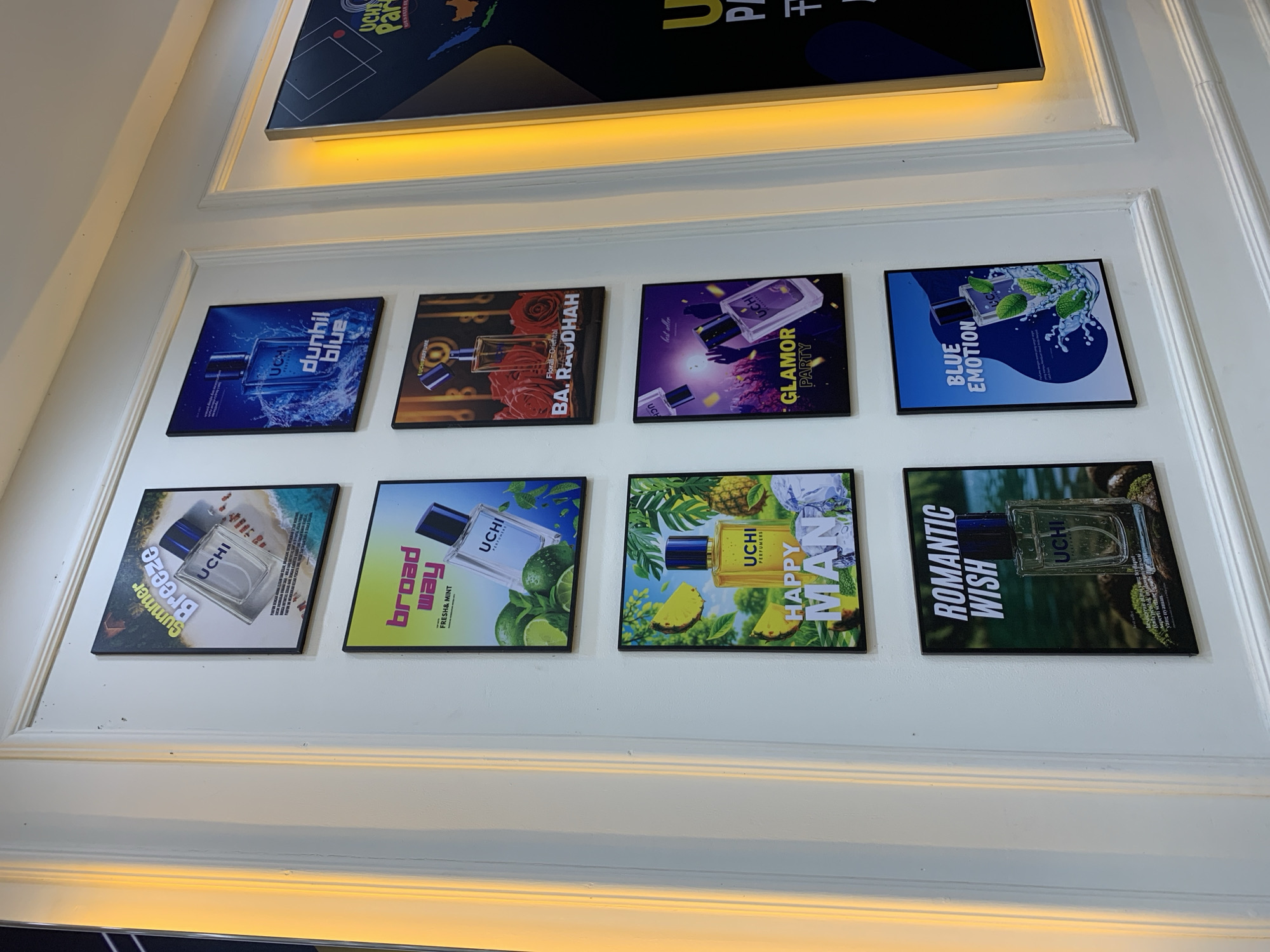 In the city of Padangjaya, Indonesia, a white, classically molded wall displays a grid of eight framed advertisements for UCHI perfume. The advertisements are arranged in two columns of four. The left column features "Summer Breeze," "Broadway Way," "Happy Man," and "Romantic Wish." The right column displays "Dunhil Blue," "Ba. Raudhah," "Glamor Party," and "Blue Emotion." A large, backlit advertisement, partially visible on the far right, appears to promote UCHI perfume in a similar style. The lighting in the scene is warm and illuminated, possibly from recessed lighting or an adjacent fixture, casting a gentle glow on the wall and advertisements.