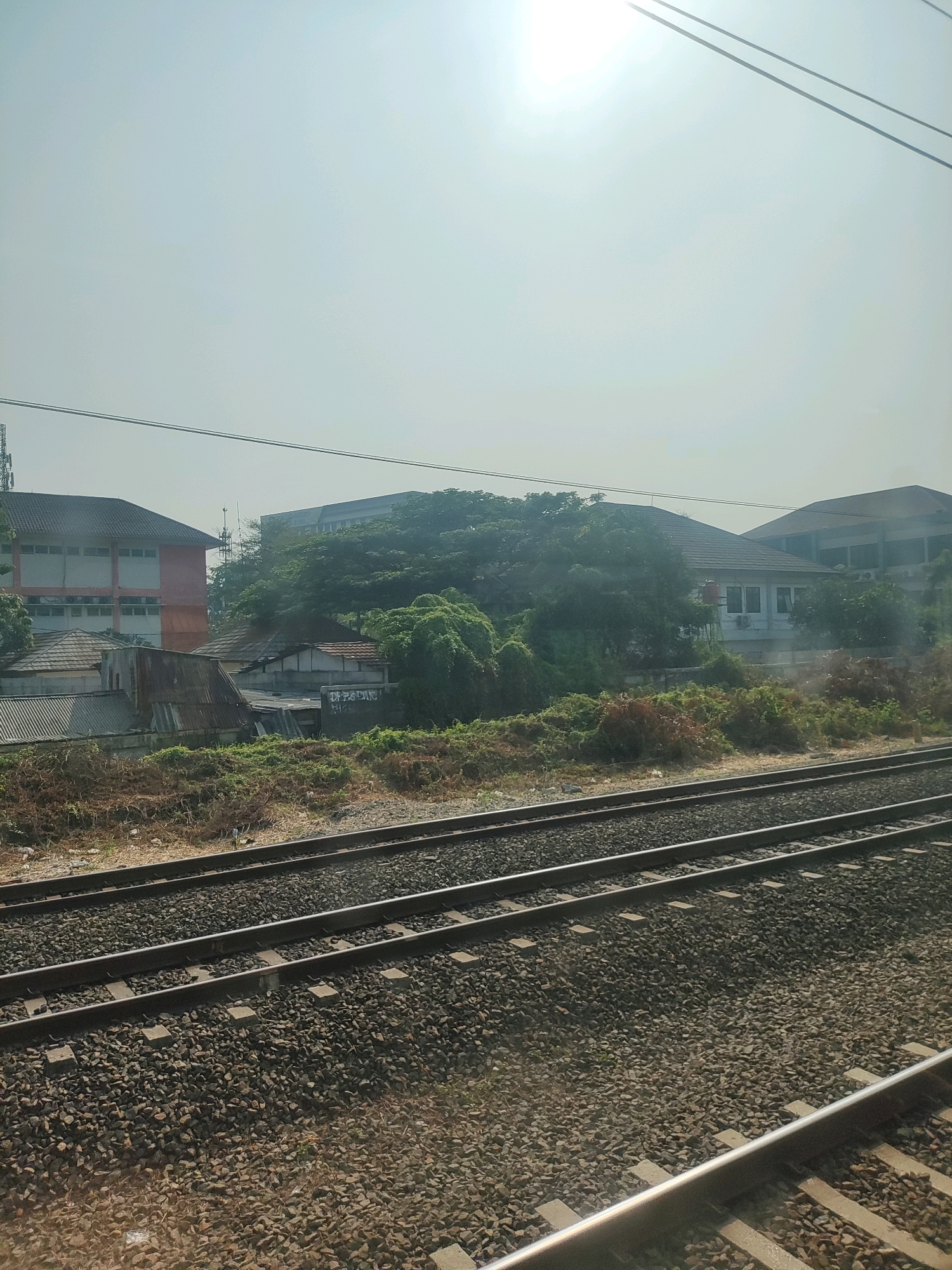 The image depicts a view of a railway line in Bekasi, Indonesia, with a bright, hazy sky overhead. The foreground is dominated by multiple sets of train tracks, densely packed with dark gravel. Beyond the tracks, a strip of dry, brown vegetation runs horizontally, transitioning into a denser area of green foliage and trees.
Several buildings are visible in the midground. To the left, a multi-story building with a reddish-orange facade and white window frames stands prominently. Adjacent to it, a cluster of smaller structures with tiled roofs and corrugated metal roofing suggests a more informal settlement. Further to the right, a single-story white building with multiple windows is partially obscured by overgrown green vines and trees. Behind this, another larger, modern-looking building with a darker facade and visible windows can be seen.
Overhead, thin power lines stretch across the frame, with one set of thicker cables running closer to the top edge. The overall impression is one of urban development adjacent to a railway corridor, with pockets of dense vegetation interspersed with residential and commercial structures. The sunlight is strong, creating a bright, washed-out effect in the sky, indicating a clear but possibly humid day.