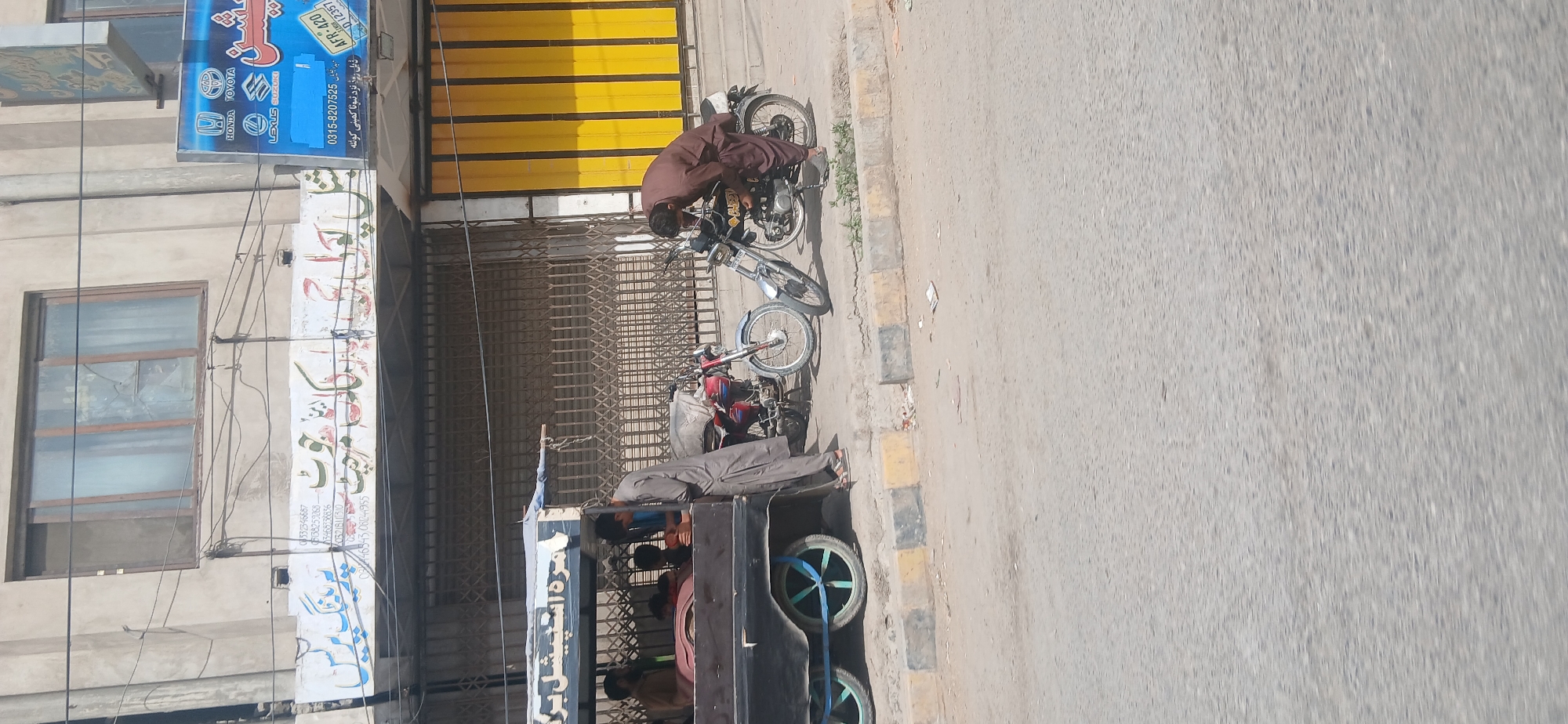 The image shows a street scene in Quetta City Tehsil, Pakistan. In the foreground, a black cart with blue-rimmed wheels is parked. Inside the cart, several people are visible, including a man in a brown shirt and others seated. A man in a grey shalwar kameez stands beside the cart, facing away from the camera.
To the right of the cart, two motorcycles are parked on the side of the road. A man in a brown shalwar kameez is bent over one of the motorcycles, appearing to be working on it. The motorcycle has a black seat and a silver fuel tank. The other motorcycle, parked behind it, is red and blue.
In the background, a building with several windows is visible. A sign with text in Arabic script is mounted above the street level. Further to the right, a bright yellow metal shuttered storefront is visible. The street itself appears to be paved, with a raised sidewalk in the middle ground. The overall scene depicts a typical urban street environment.